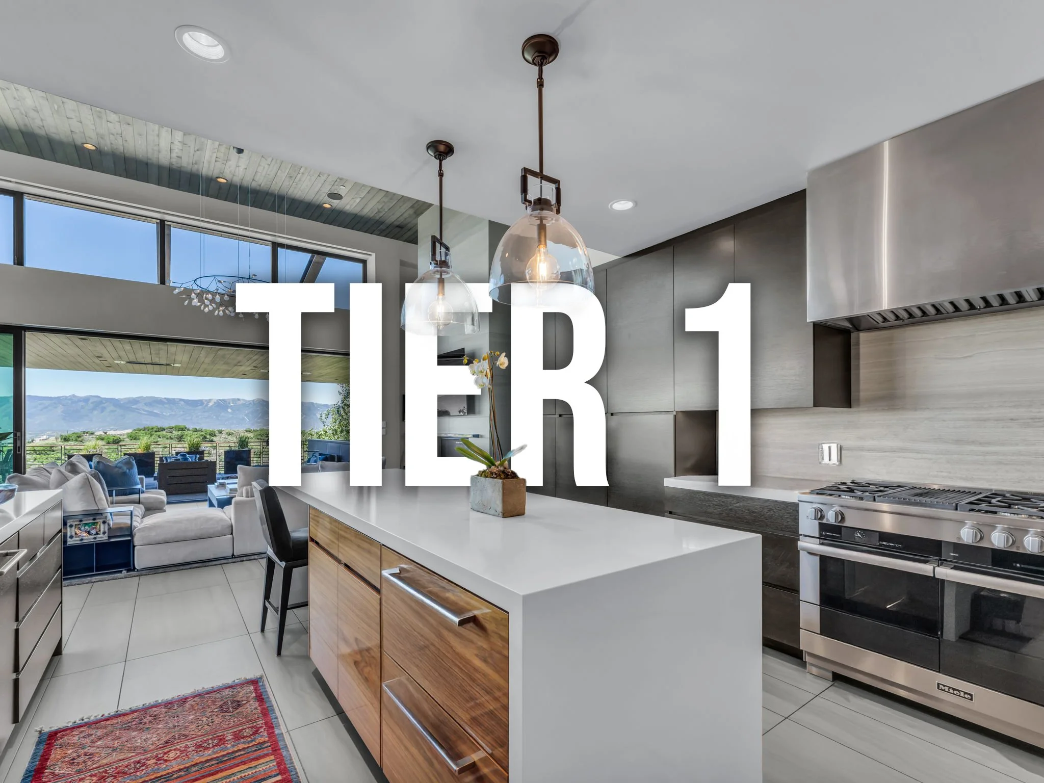 Modern kitchen with island, pendant lighting, stainless steel oven, and large windows overlooking a patio with mountains in the background.