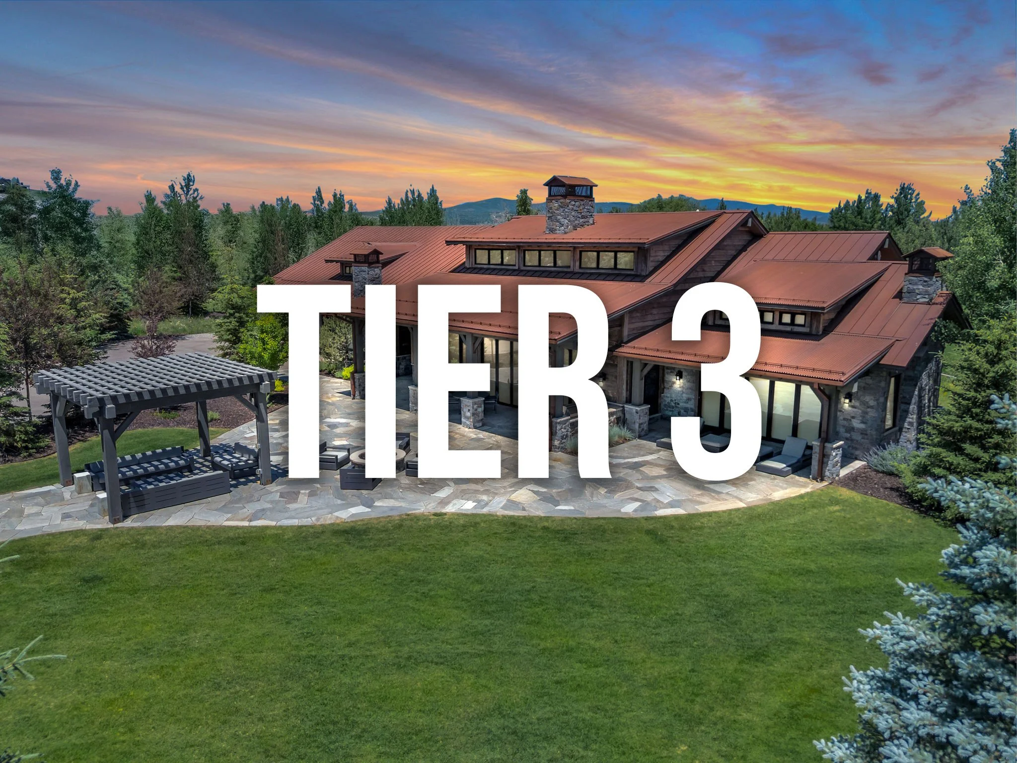 A large house with a reddish-brown metal roof, stone walls, and large windows, under a colorful sunset sky, surrounded by trees and a green lawn with a stone patio and a pergola.