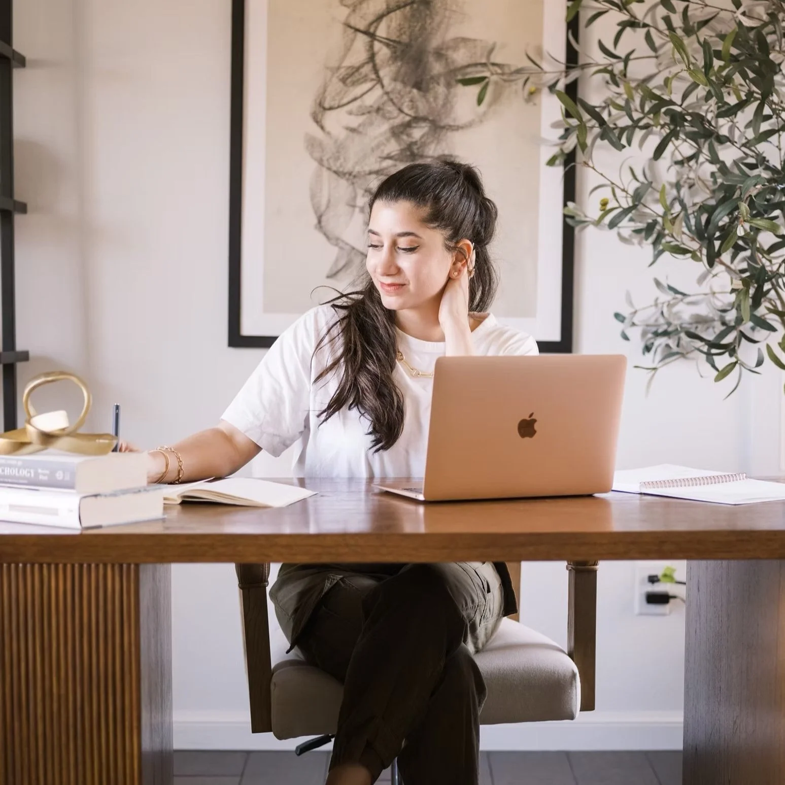 Person sitting at a desk with a laptop, writing in a notebook, surrounded by books and a plant, in a modern office setting.