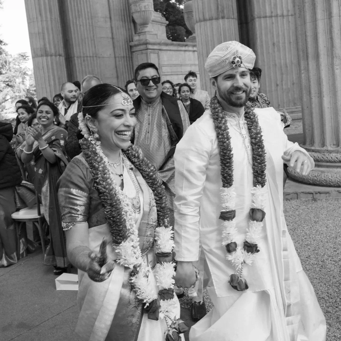 Bride and groom in traditional South Indian wedding attire walk hand in hand, smiling and wearing floral garlands, as guests applaud during an outdoor ceremony at a grand columned venue.