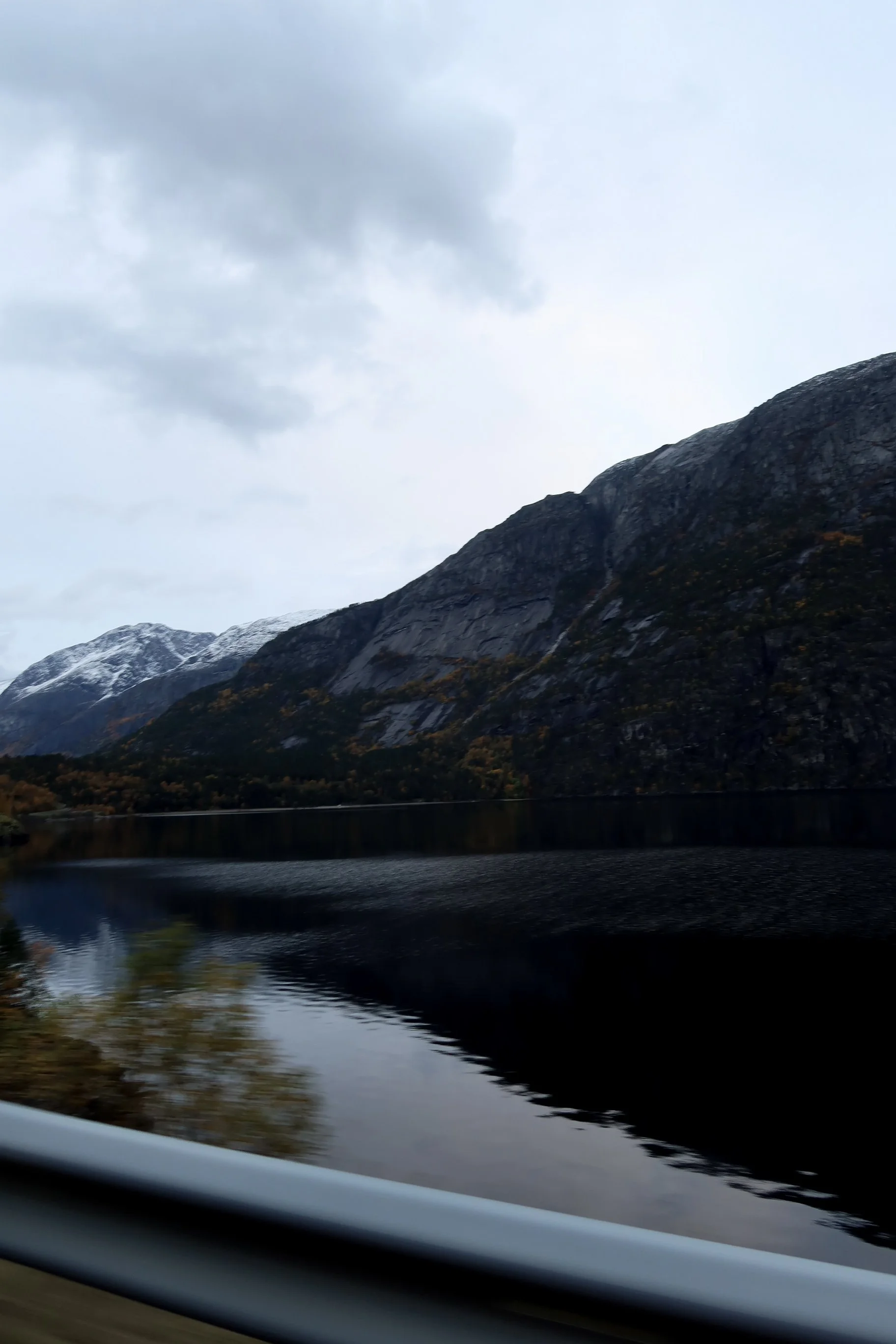 Scenic view of mountains with snow-capped peaks and a calm lake reflecting the mountains and cloudy sky, taken from a moving vehicle.