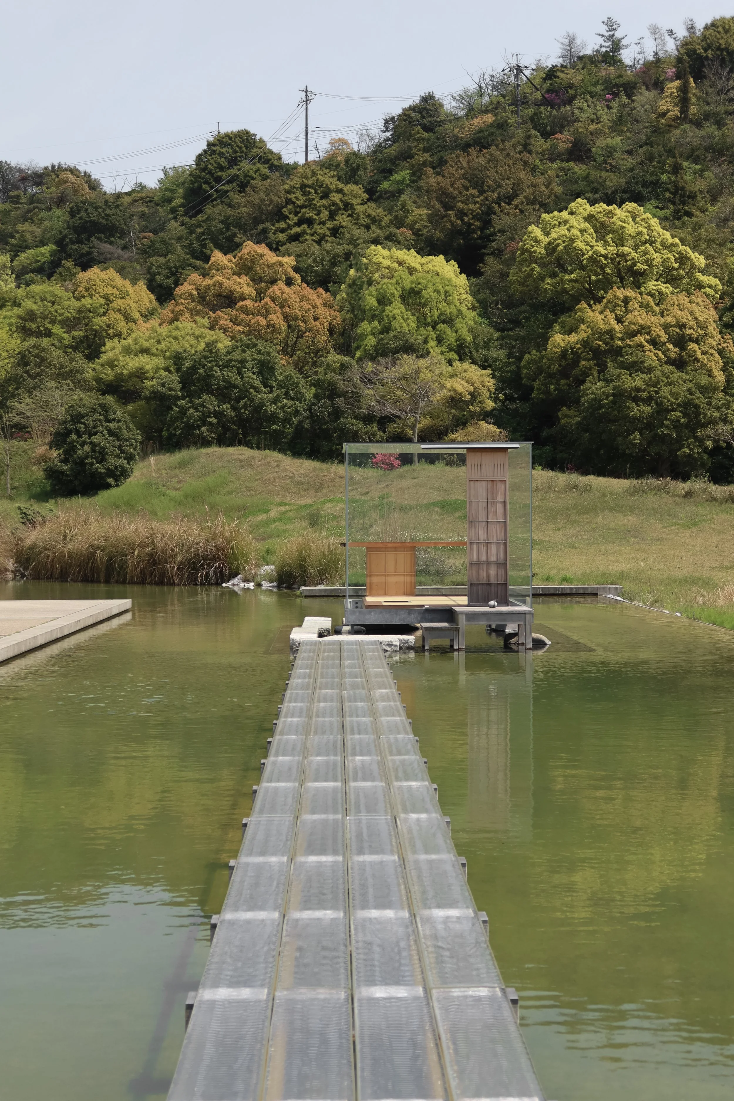 A pathway extends over water towards a small wooden structure with a glass wall, with a lush green hillside in the background.