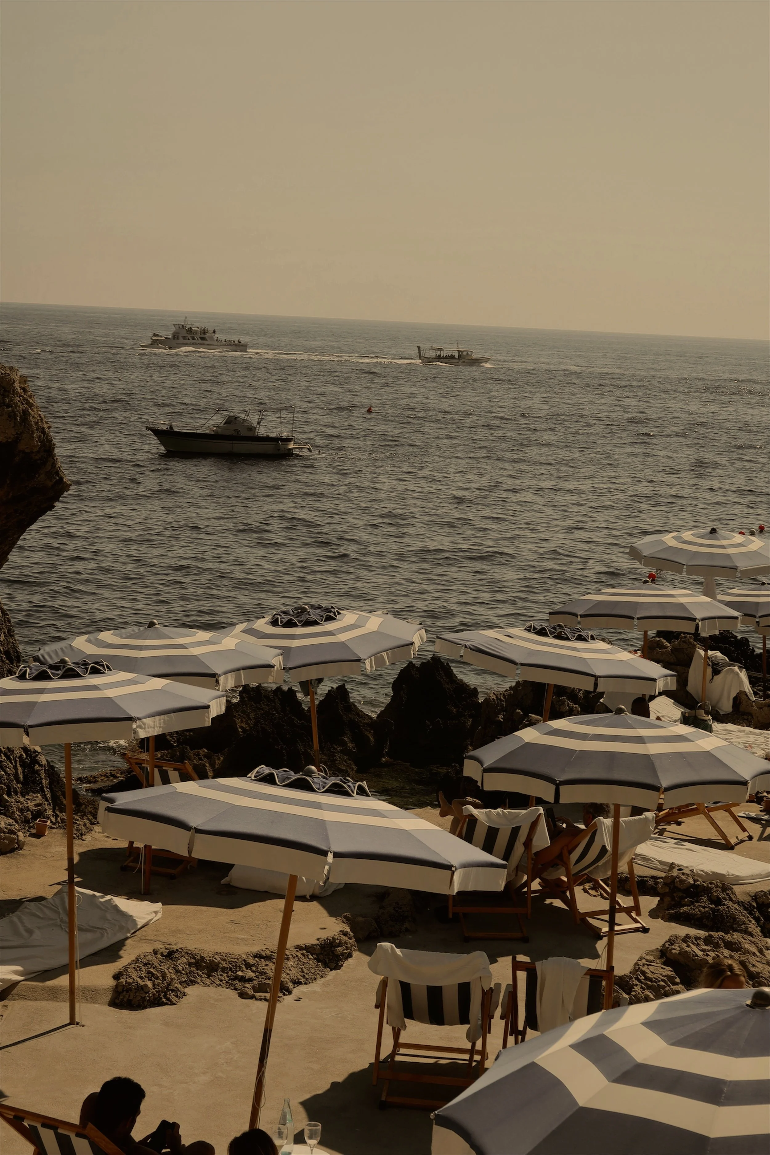 Beach with striped umbrellas, lounge chairs, and rocks along the shore, with boats sailing on the ocean in the background.