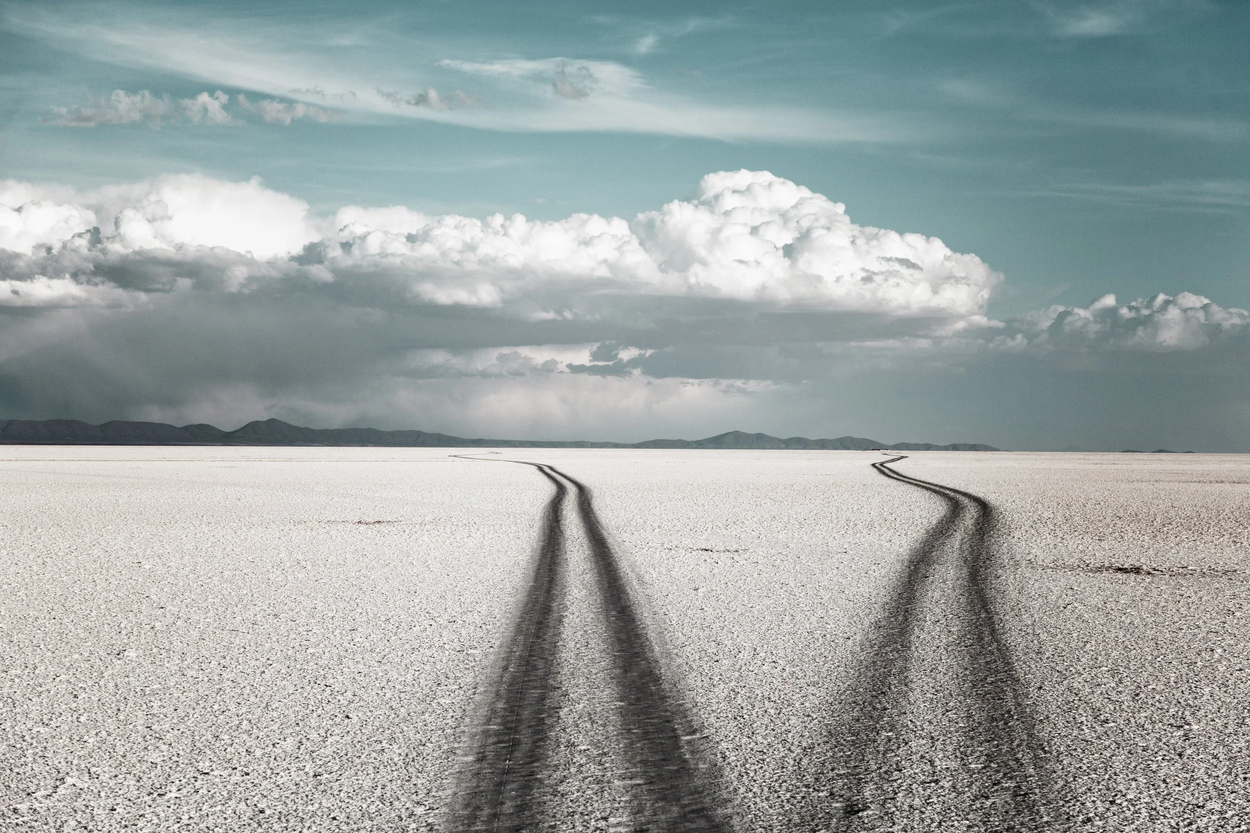 Two tire tracks on a vast, flat, white salt flat with a mountain range in the distance and a partly cloudy sky.