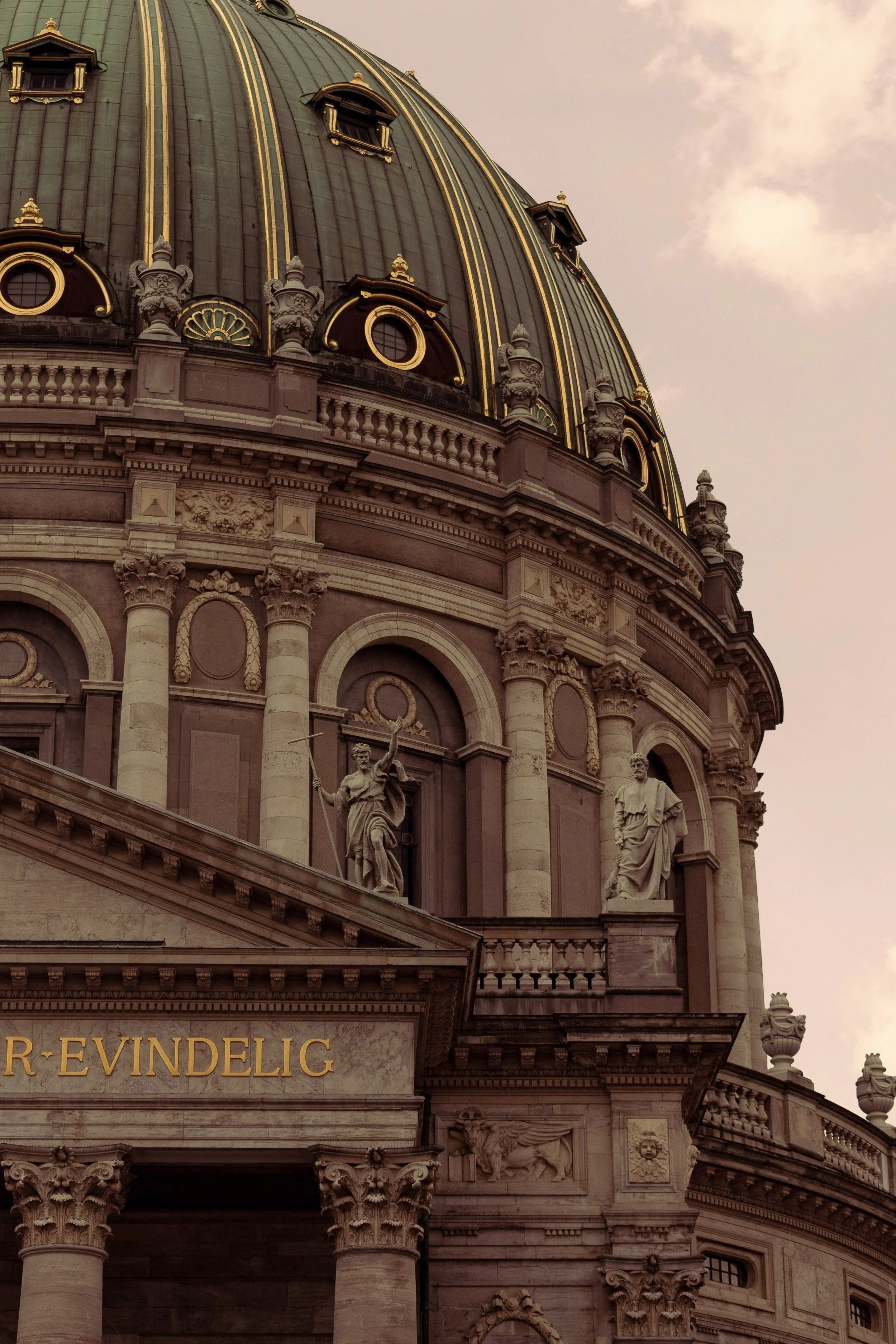 Close-up of a historical building's ornate facade with statues and gold accents on a domed roof, under a cloudy sky.