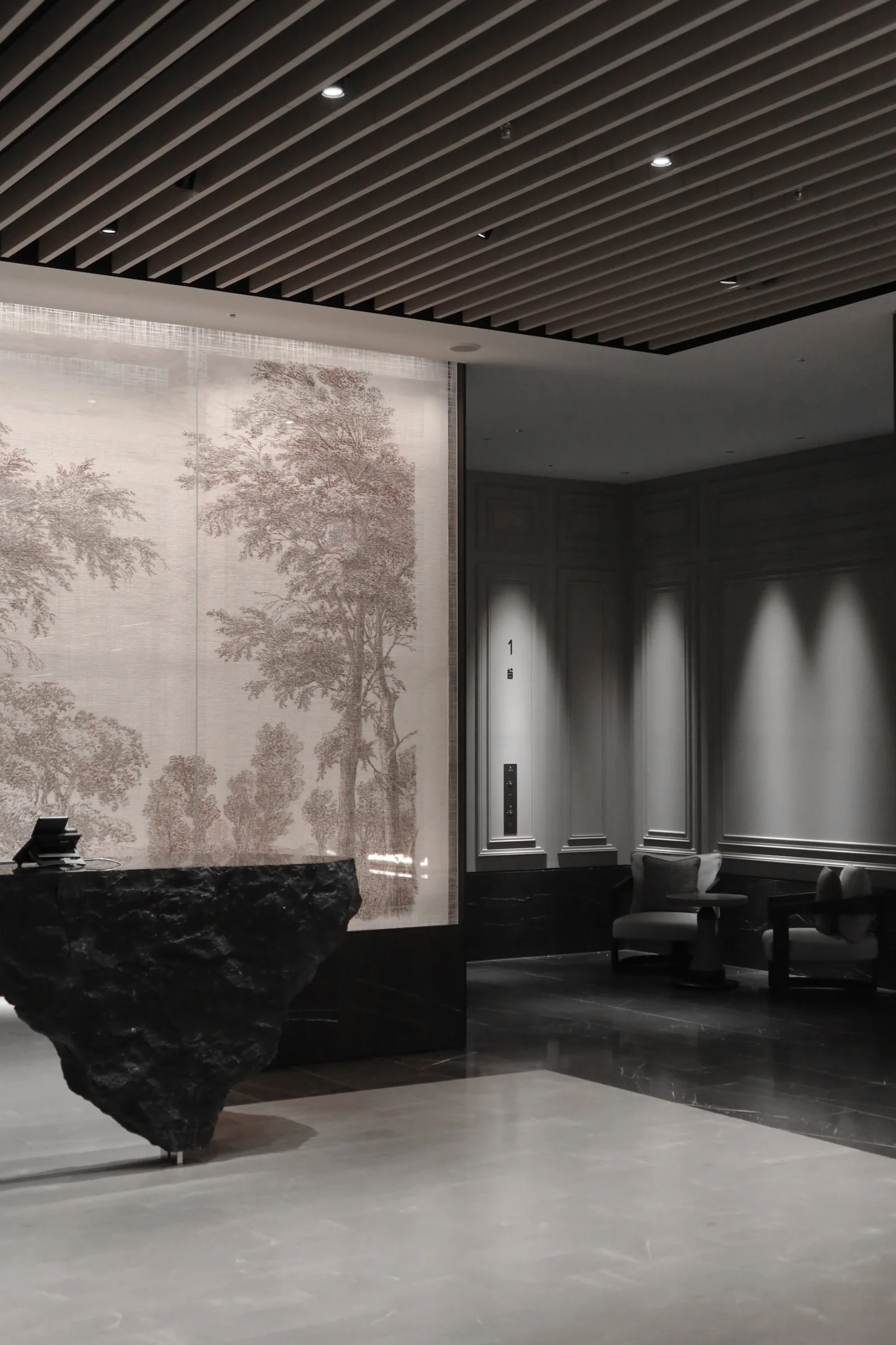 Modern hotel lobby with a black marble reception desk, a large wall mural of trees, seating area with dark gray chairs, and elevator with a minimalistic design