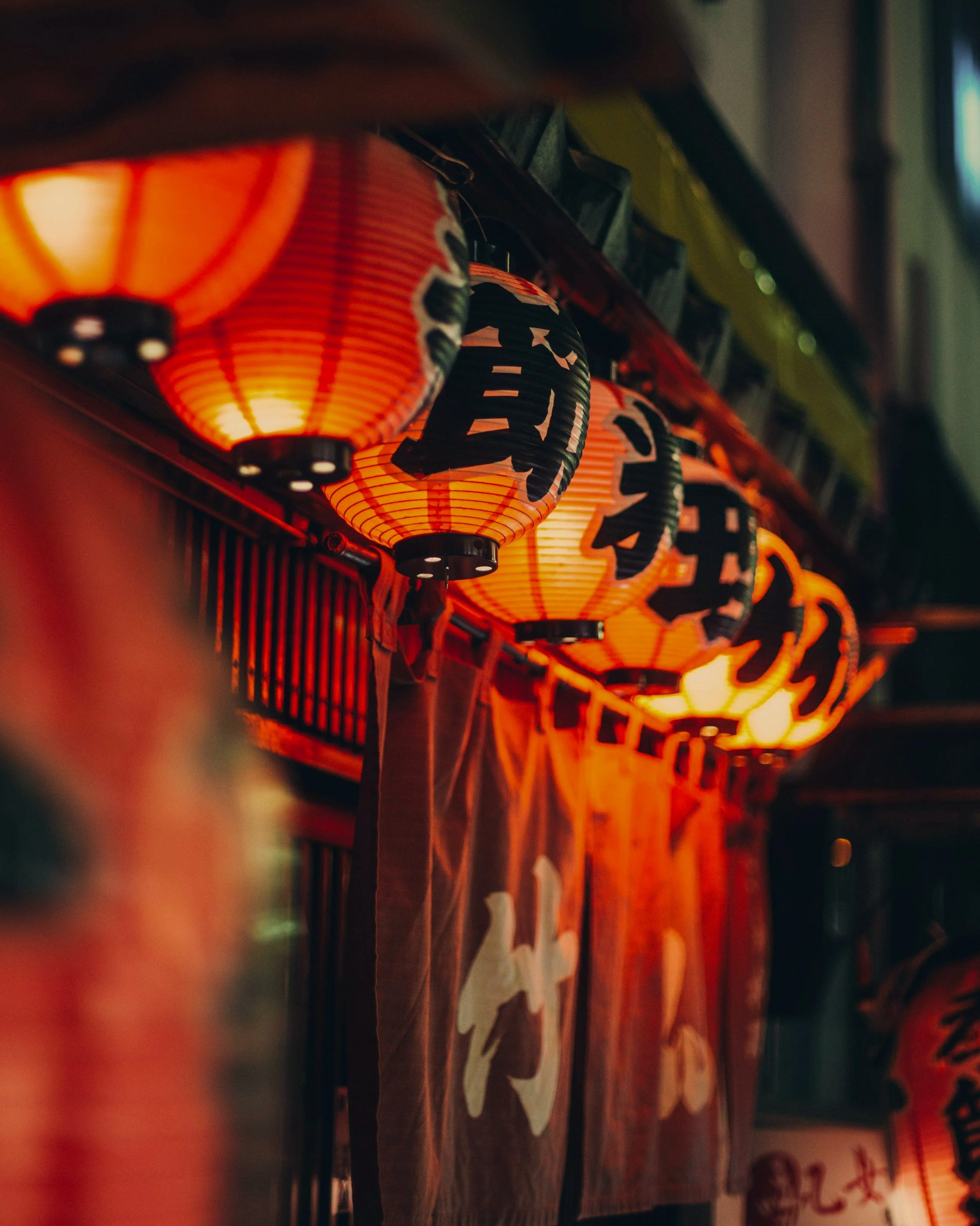 A row of illuminated Japanese paper lanterns hanging at night, with Japanese characters written on them and fabric banners visible beneath.