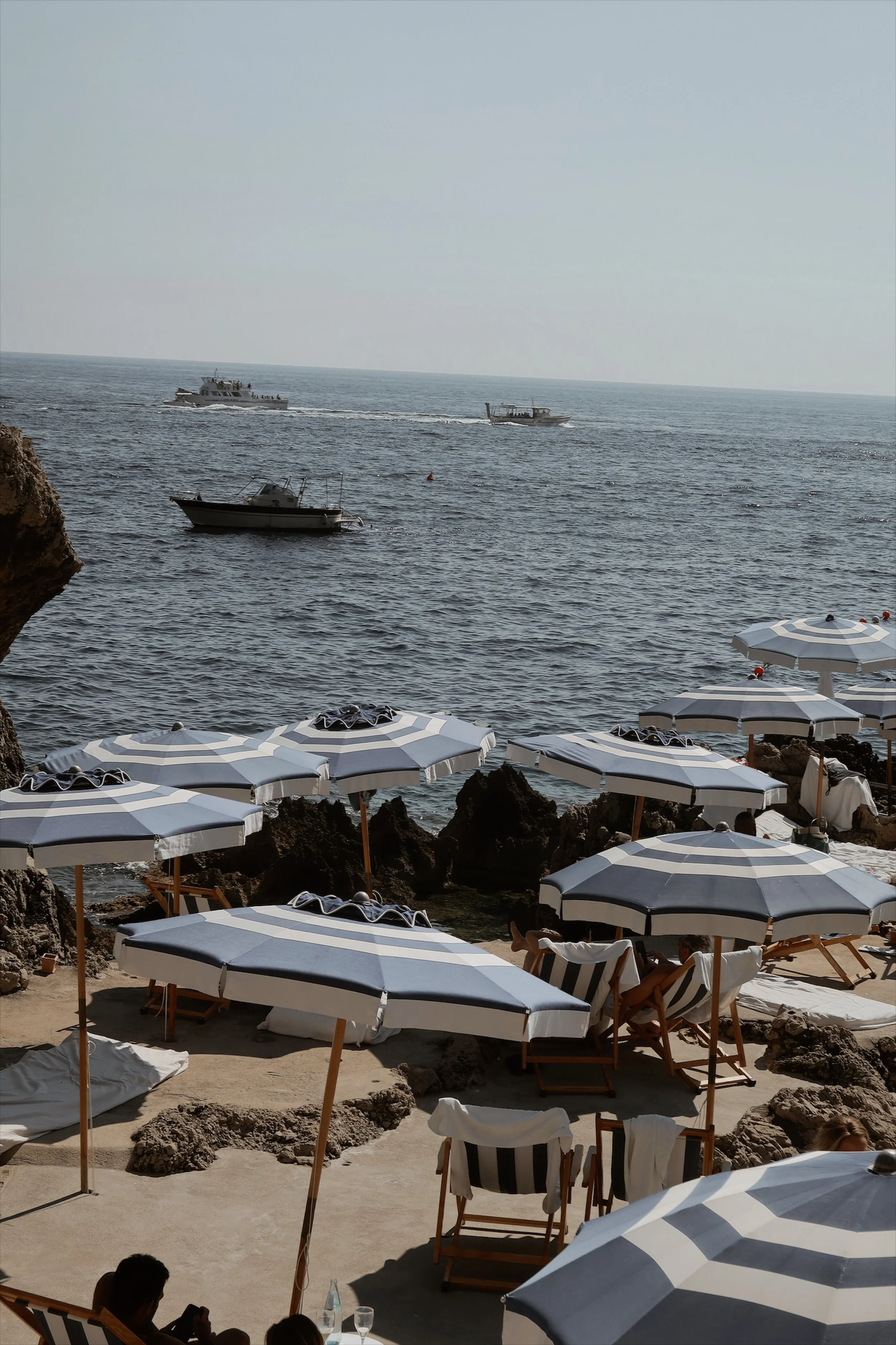Beach scene with striped umbrellas, lounge chairs, rocks, and boats on the water in the background.