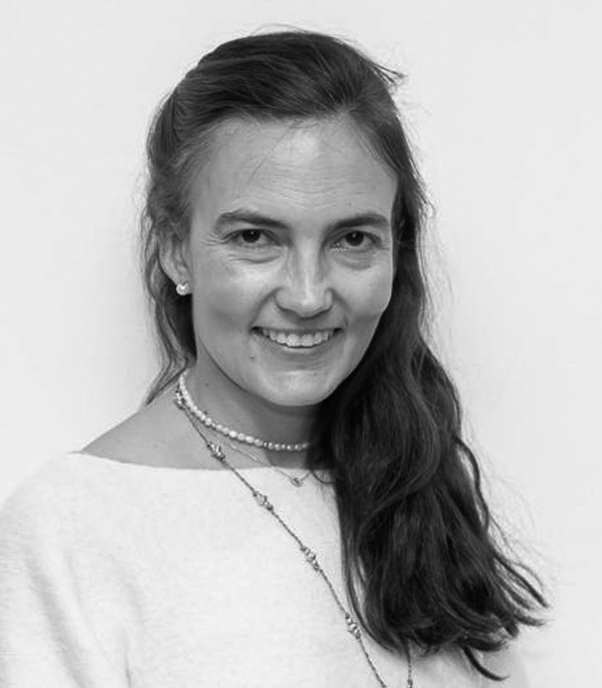 Black and white portrait of a smiling woman with long, dark hair, wearing earrings, layered necklaces, and a light-colored top.