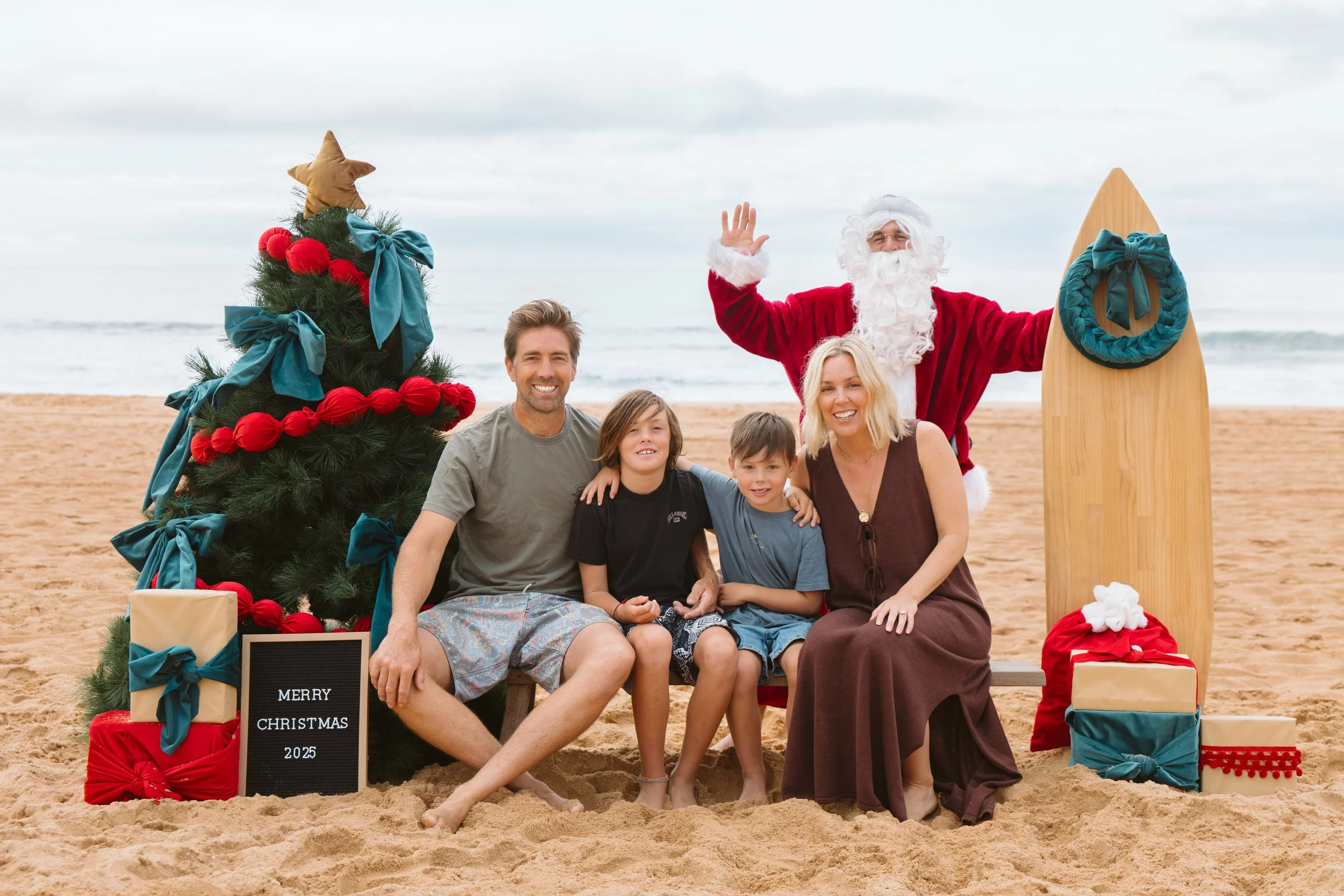 Santa Photos Narrabeen Beach. A family sitting on the beach with a decorated Christmas tree, Santa Claus, and a surfboard, celebrating Christmas in 2025.