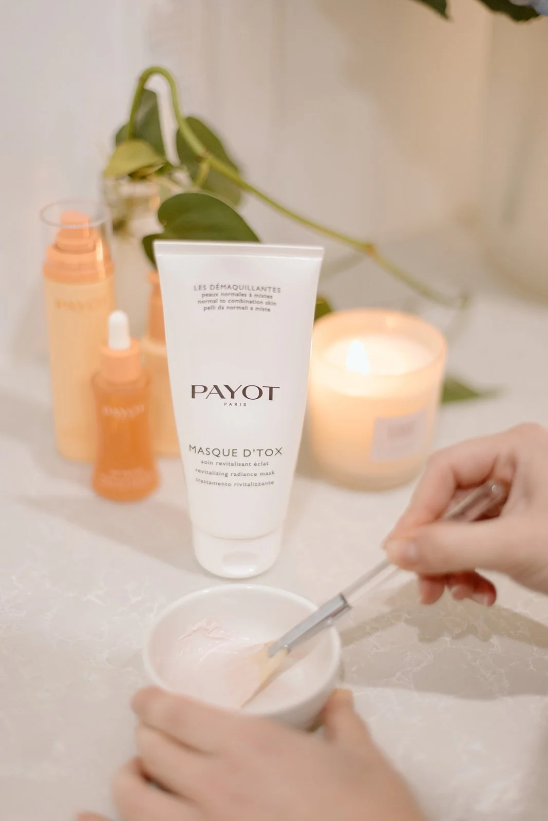 A person mixing a skincare product in a small bowl with a metal spatula, with various skincare bottles and a lit candle on a white surface in the background.