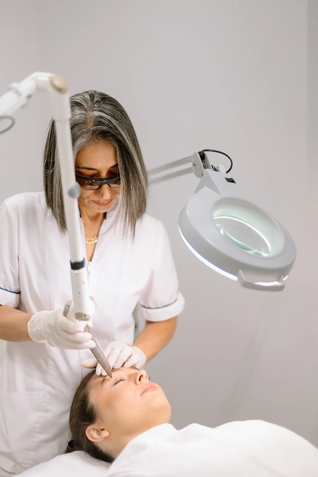 A woman in a white medical coat and gloves is performing a cosmetic procedure on a woman's face in a clinical setting, using a handheld device under a magnifying light.