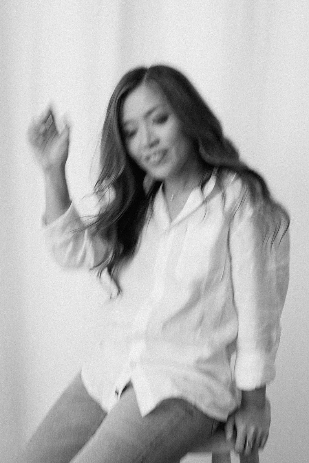 Black and white photograph of a woman with long, dark hair, wearing a white shirt and jeans, sitting on a stool against a plain wall. The photo is intentionally blurry.