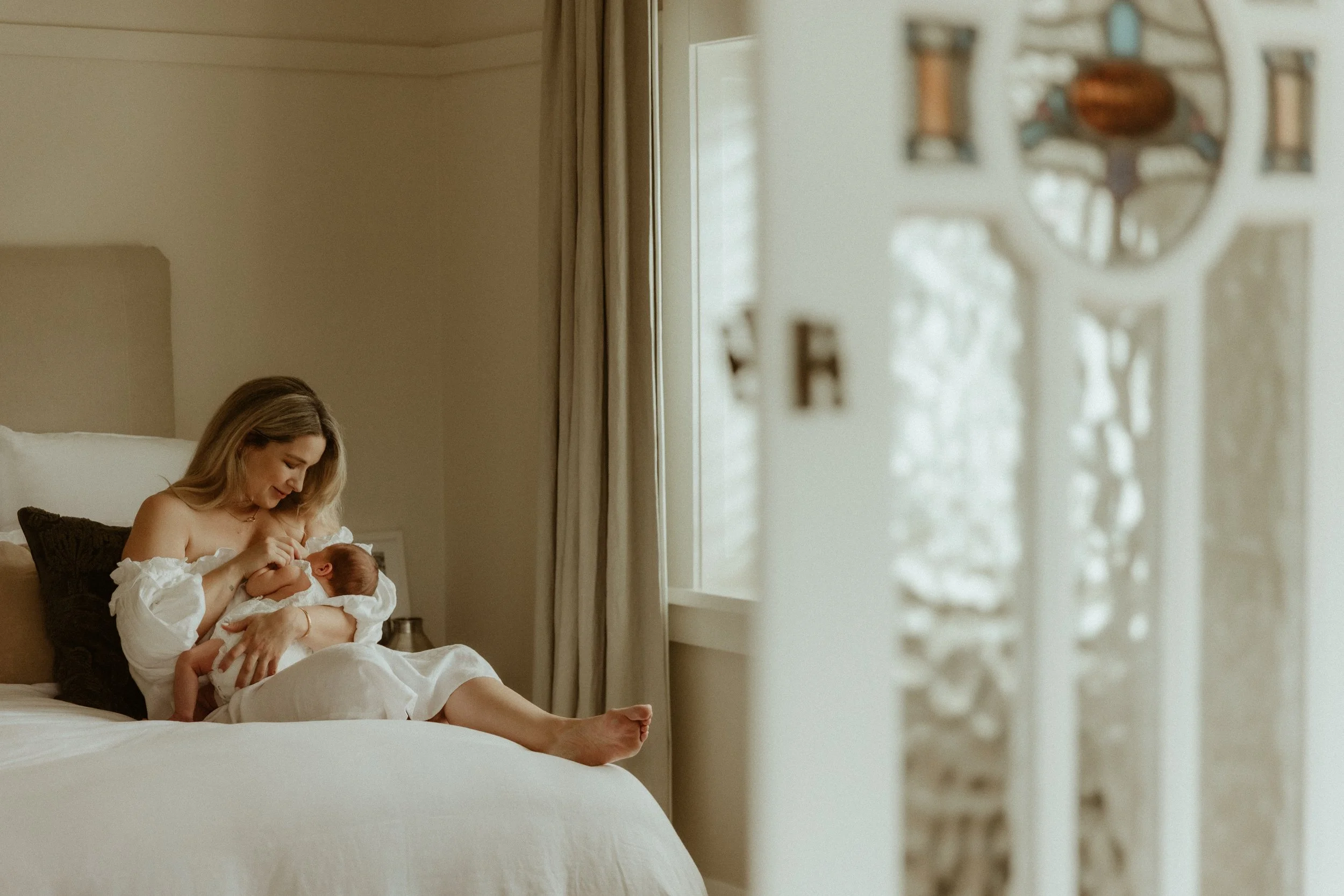 A woman sitting on a bed, breastfeeding and looking at her newborn baby indoors with sunlight coming through a window.