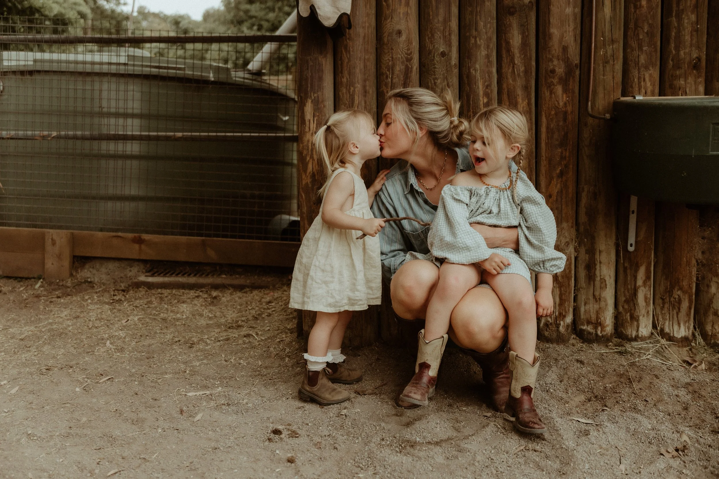 A woman and two young girls with blonde hair sitting against a wooden structure outdoors, with one girl giving a kiss on the lips to the woman and the other girl smiling and looking on. The scene appears rustic and natural, suggesting a farm or rural