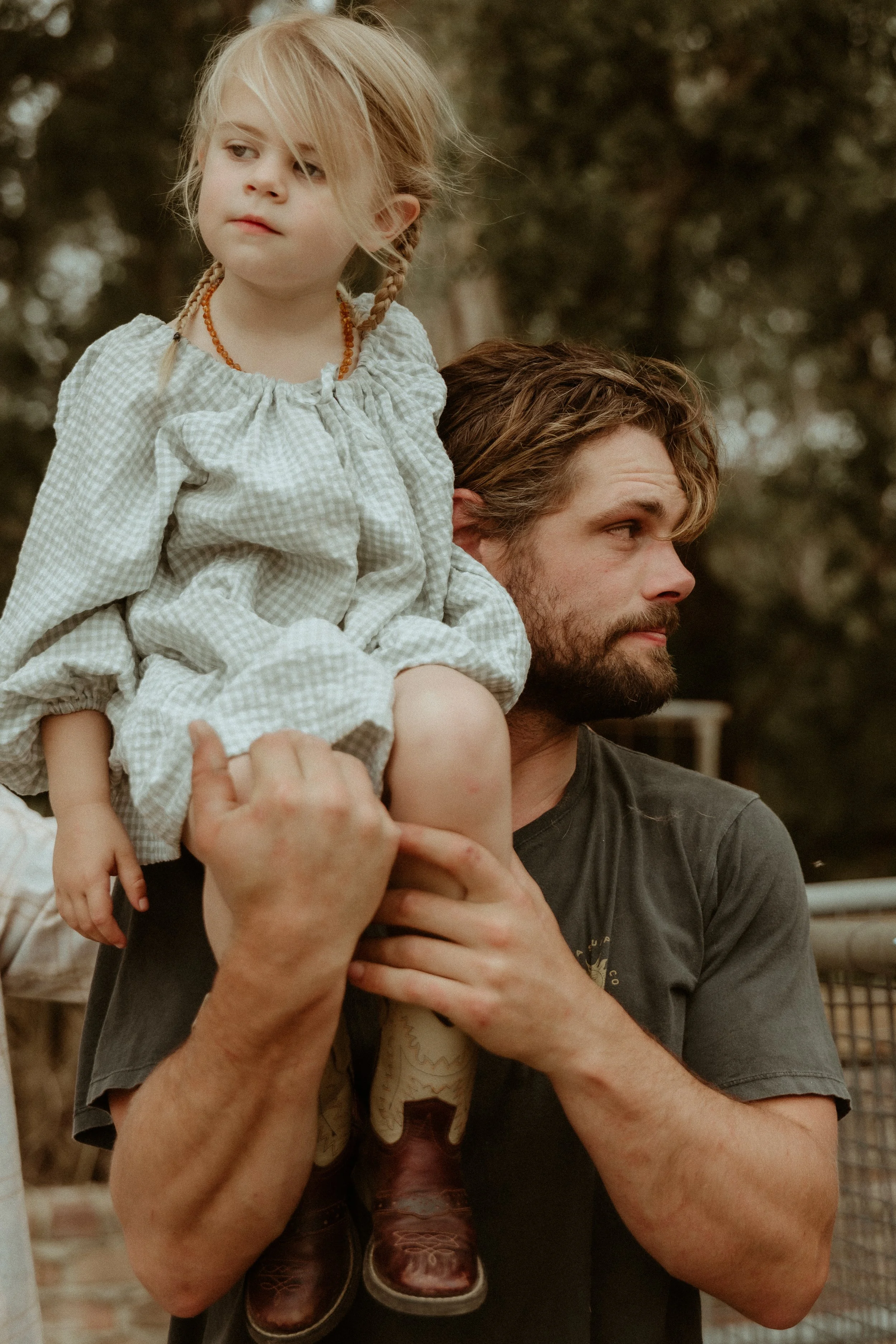 A man with a beard carries a young girl on his shoulder outdoors. The girl has blonde hair styled in braids and is wearing a light-colored gingham dress. The man has short, wavy hair and is looking to the side. Trees and fencing are visible in the ba