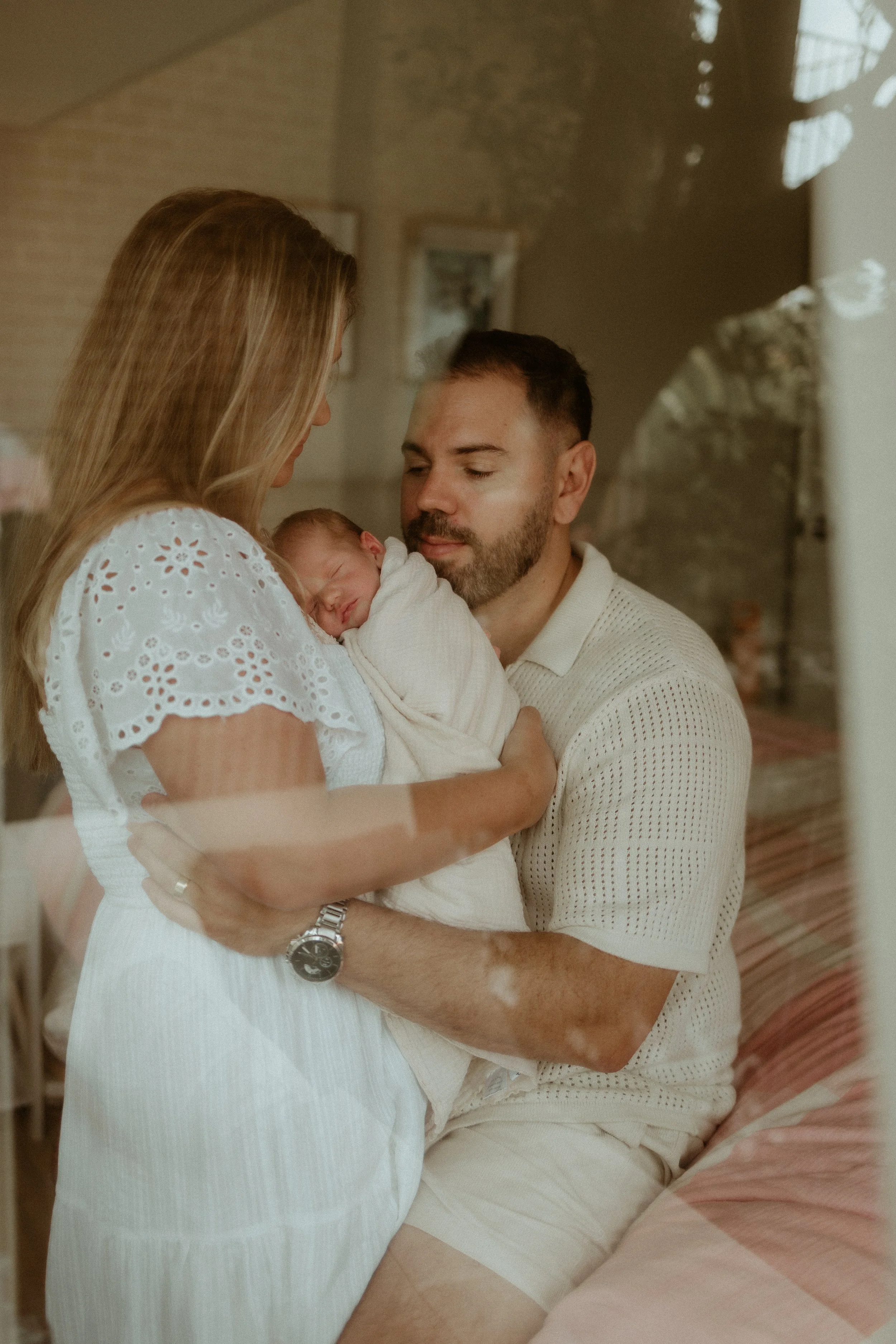 A woman and a man holding an infant, with the woman holding the baby close to her chest and the man looking at the baby. They are indoors and appear to be a family moment.
