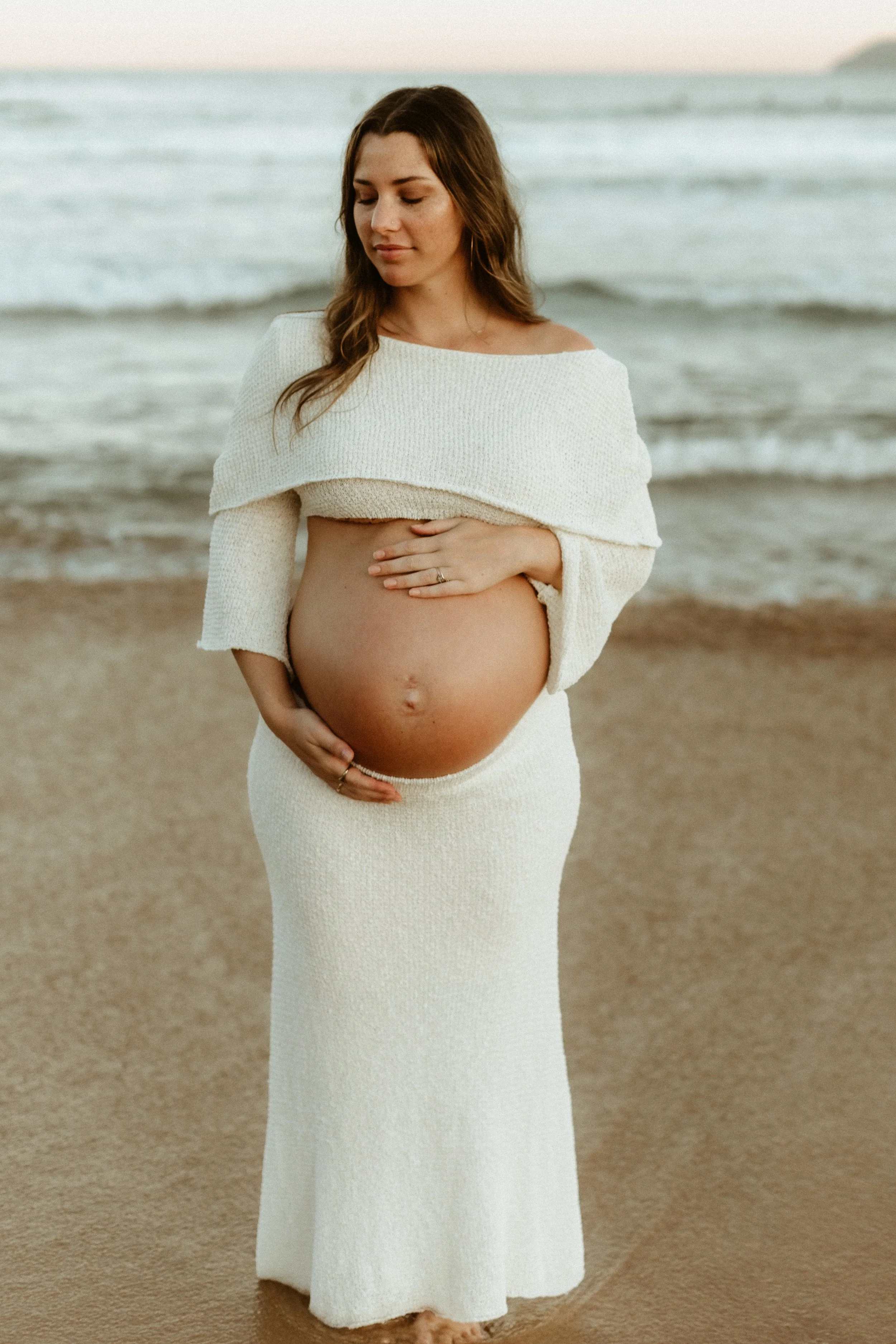 Pregnant woman standing on the beach in a white knit dress, gently holding her belly, with the ocean and a distant landmass in the background.