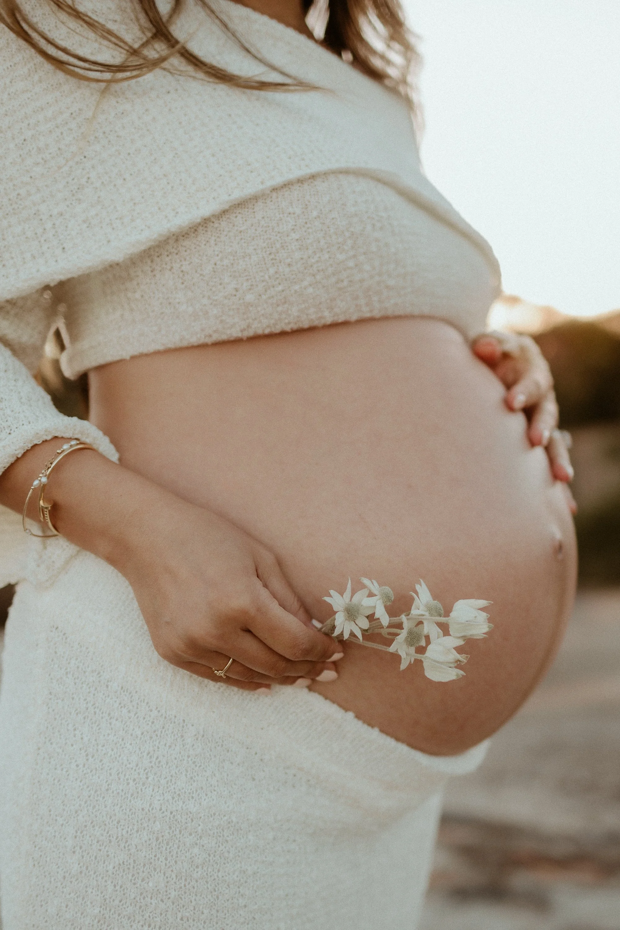A pregnant woman holding a small bouquet of white flowers on her belly, dressed in a light-colored, textured outfit, with her hand gently resting on her abdomen.