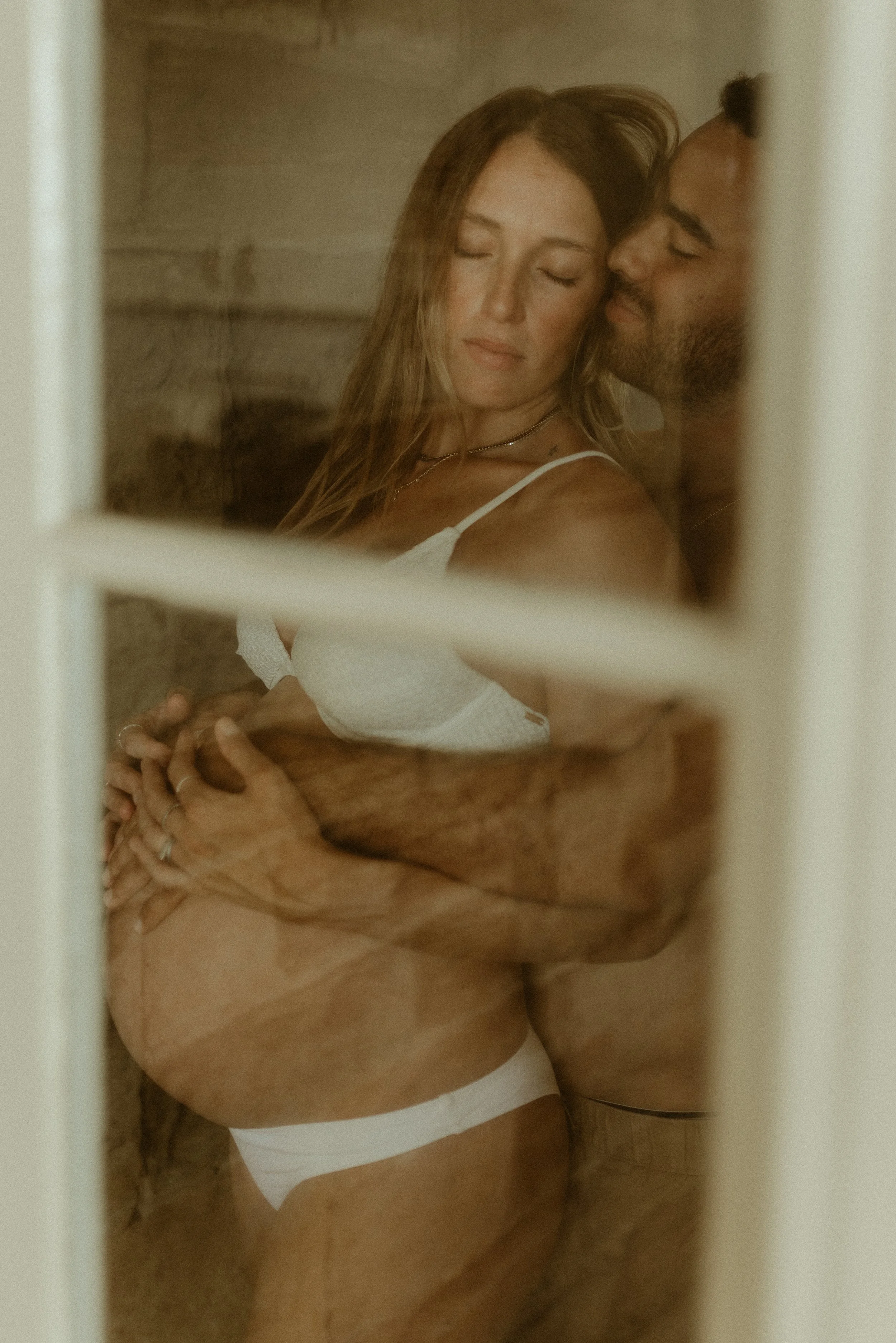 A man and woman embracing closely with eyes closed through a window or glass panel.
