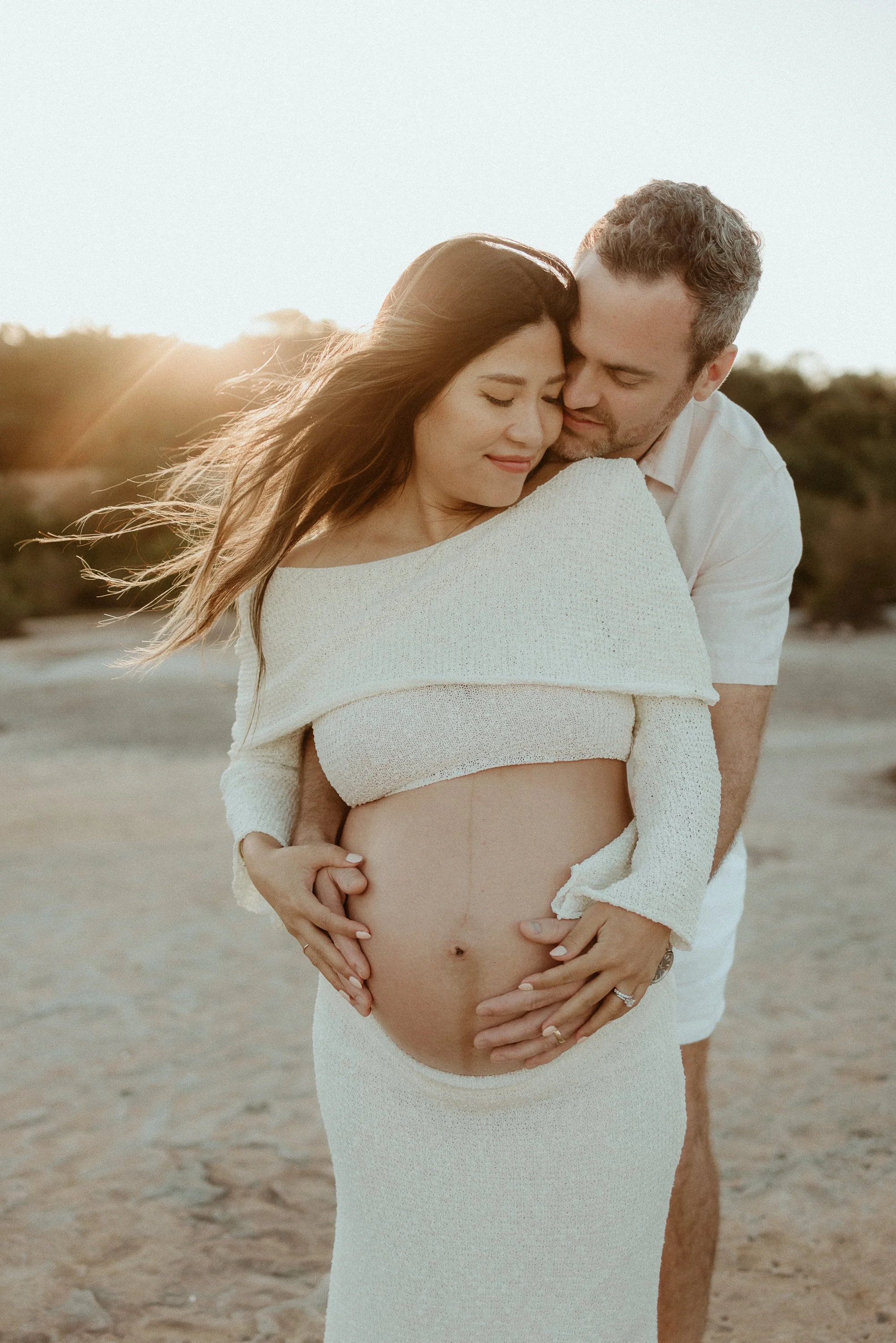 A pregnant woman and a man sharing an intimate moment on a beach at sunset, with the man holding the woman's pregnant belly.