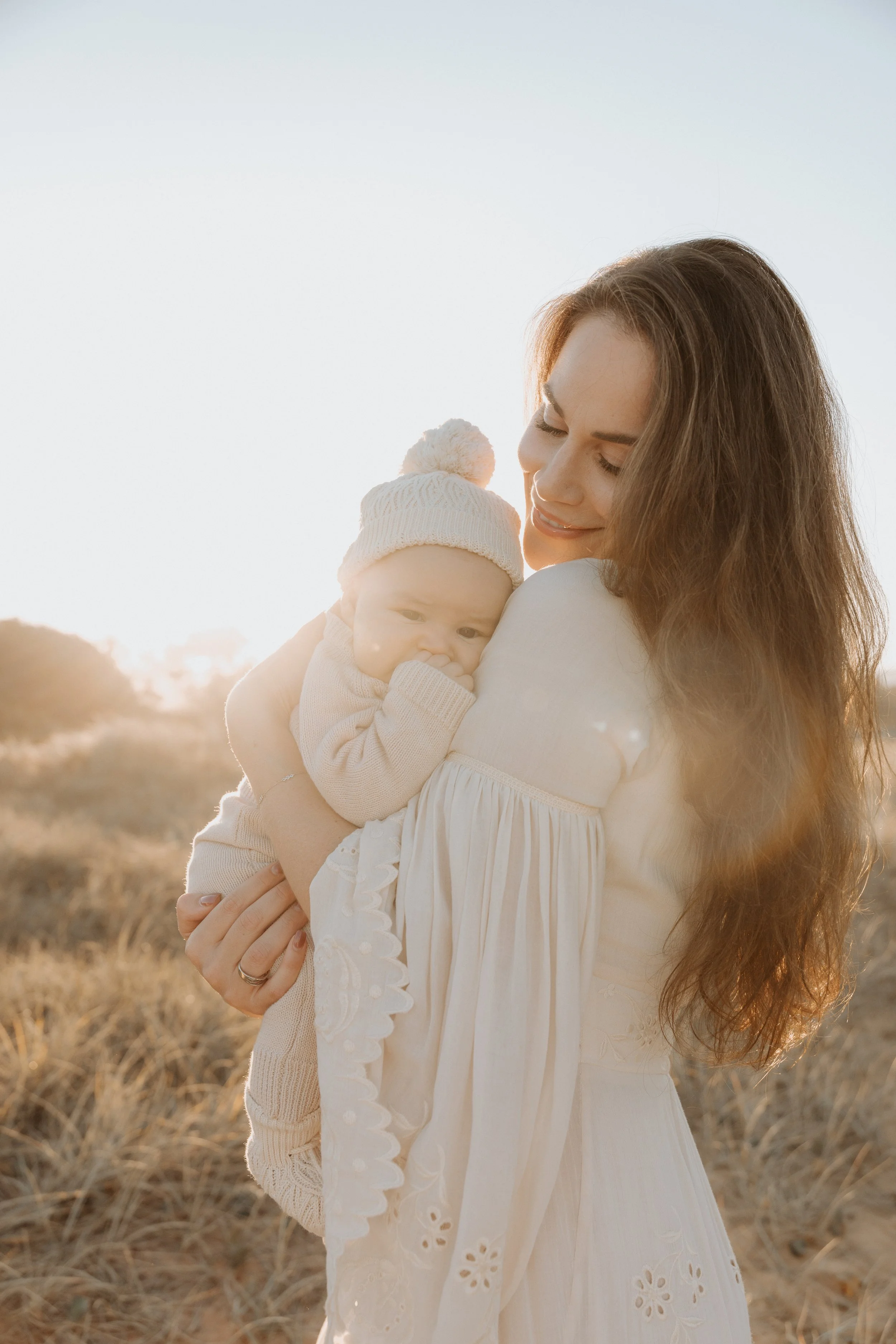 A woman holding a baby on a sunny outdoor field during sunset, both dressed in light-colored clothing.