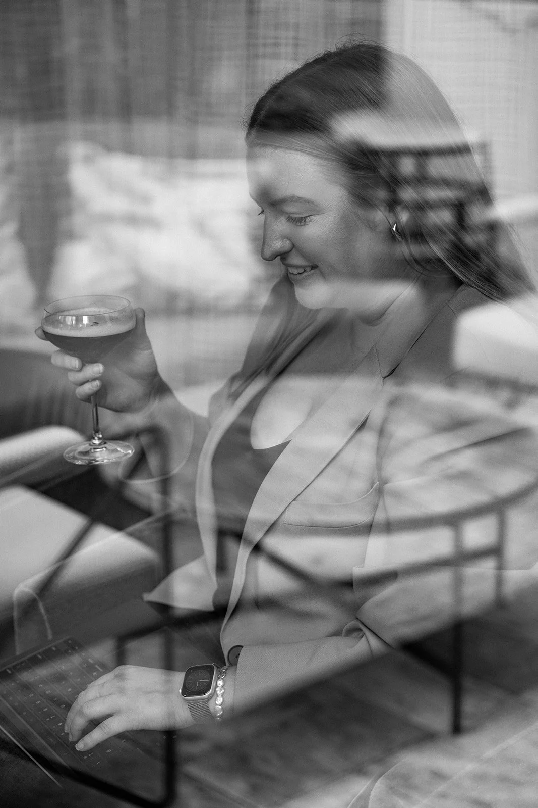 A woman with long hair, wearing a blazer and watch, sitting at a desk, holding a glass of wine and working on a laptop, seen through a window with reflections.
