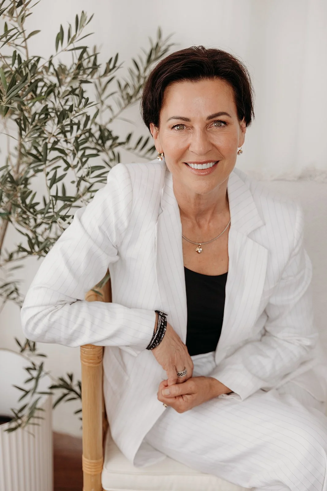 A smiling woman with short dark hair, wearing a white pinstripe blazer and black top, seated on a wooden chair with a green potted plant in the background.