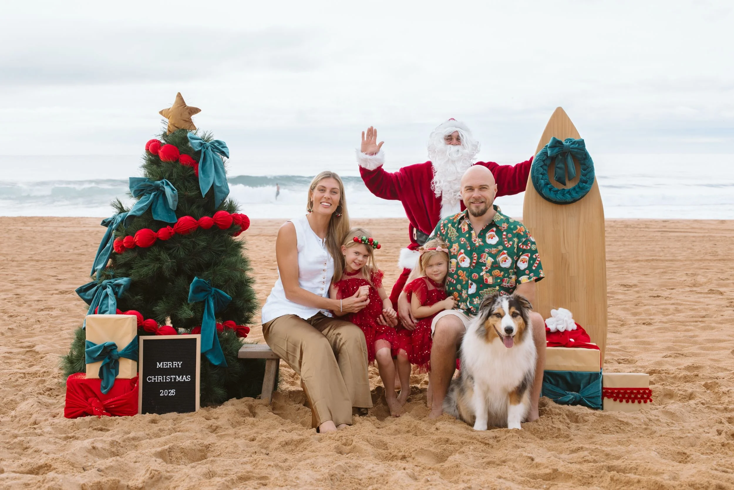 Santa Photos Narrabeen. Family celebrating Christmas on the beach with Santa Claus, a decorated Christmas tree, their dog and presents.