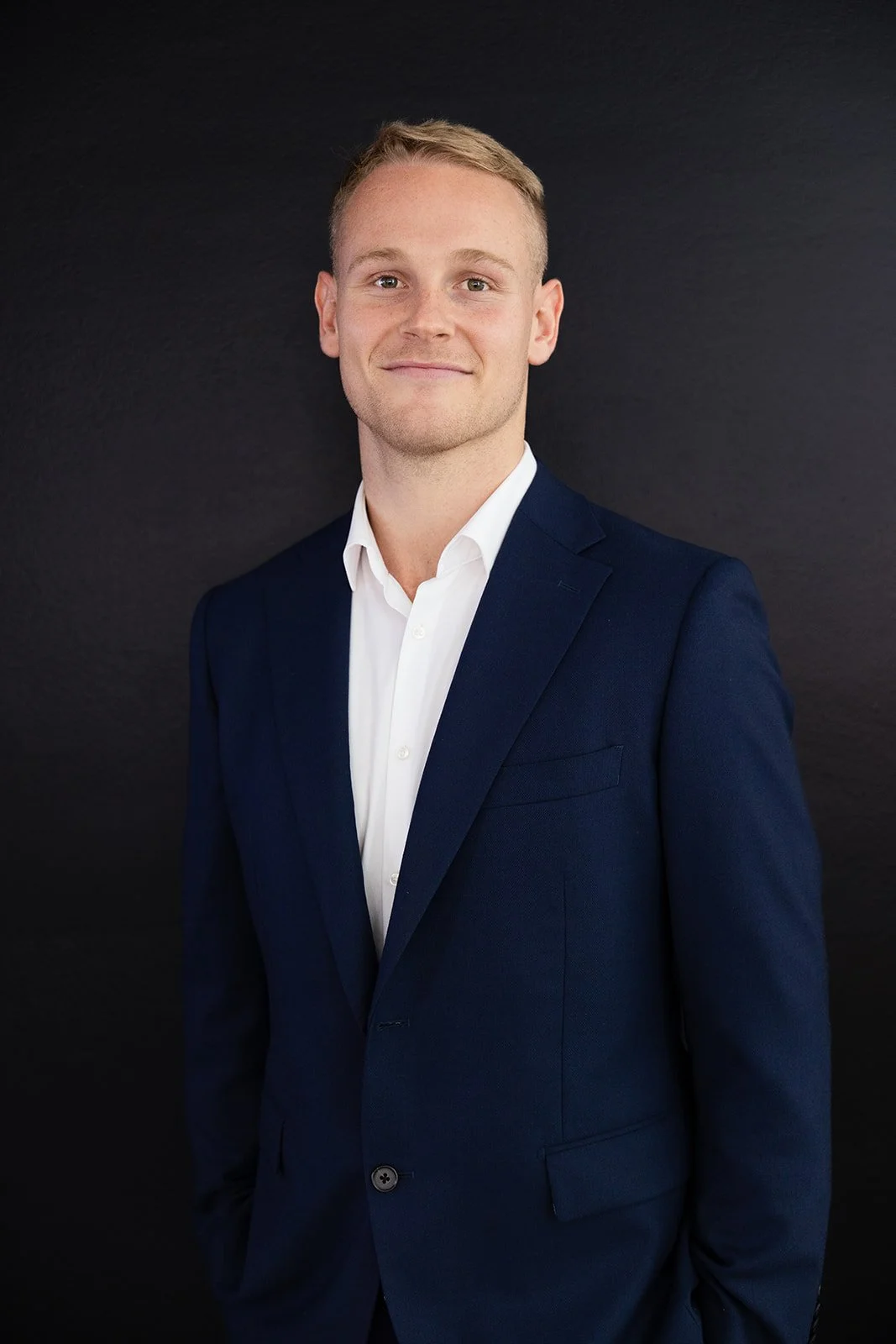 A young man with short, light-colored hair, dressed in a navy blue suit and white shirt, standing in front of a dark background.