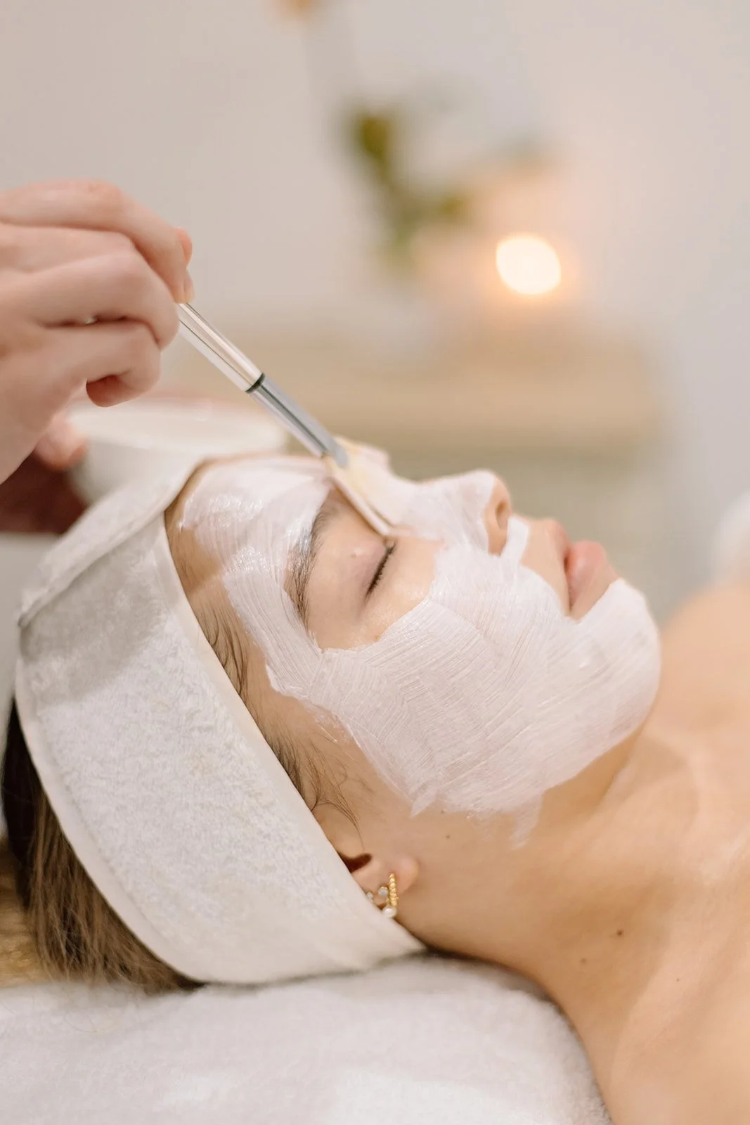 A woman is receiving a facial treatment, with her face covered in a white facial mask. She is lying down with a headband, and a skincare specialist is applying the mask with a brush.