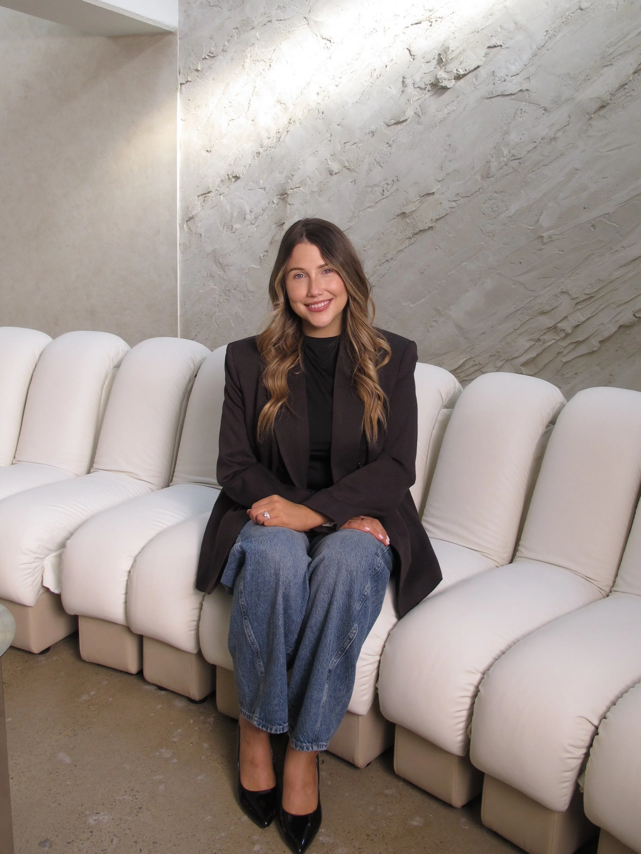 A woman sitting on a white curved sofa in front of a textured stone wall, smiling at the camera, wearing a black blazer, jeans, and black heels.
