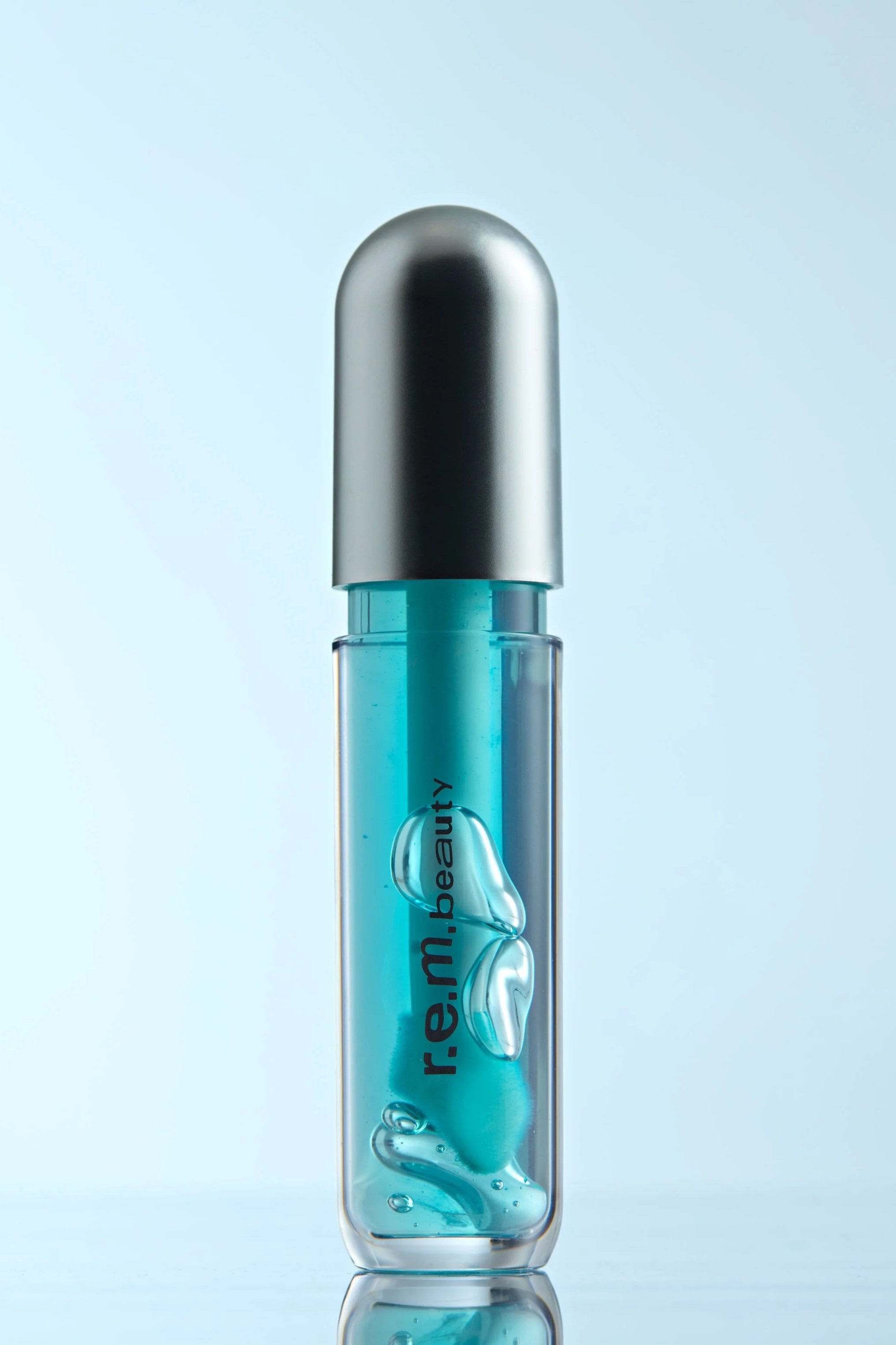 Olio labbra REM beauty, confezione trasparente con all'interno olio labbra color azzurro, poggia su base specchiata, sullo sfondo color ghiaccio, composizione luminosa e moderna che valorizza texture glossy, colore e finish nutriente del prodotto