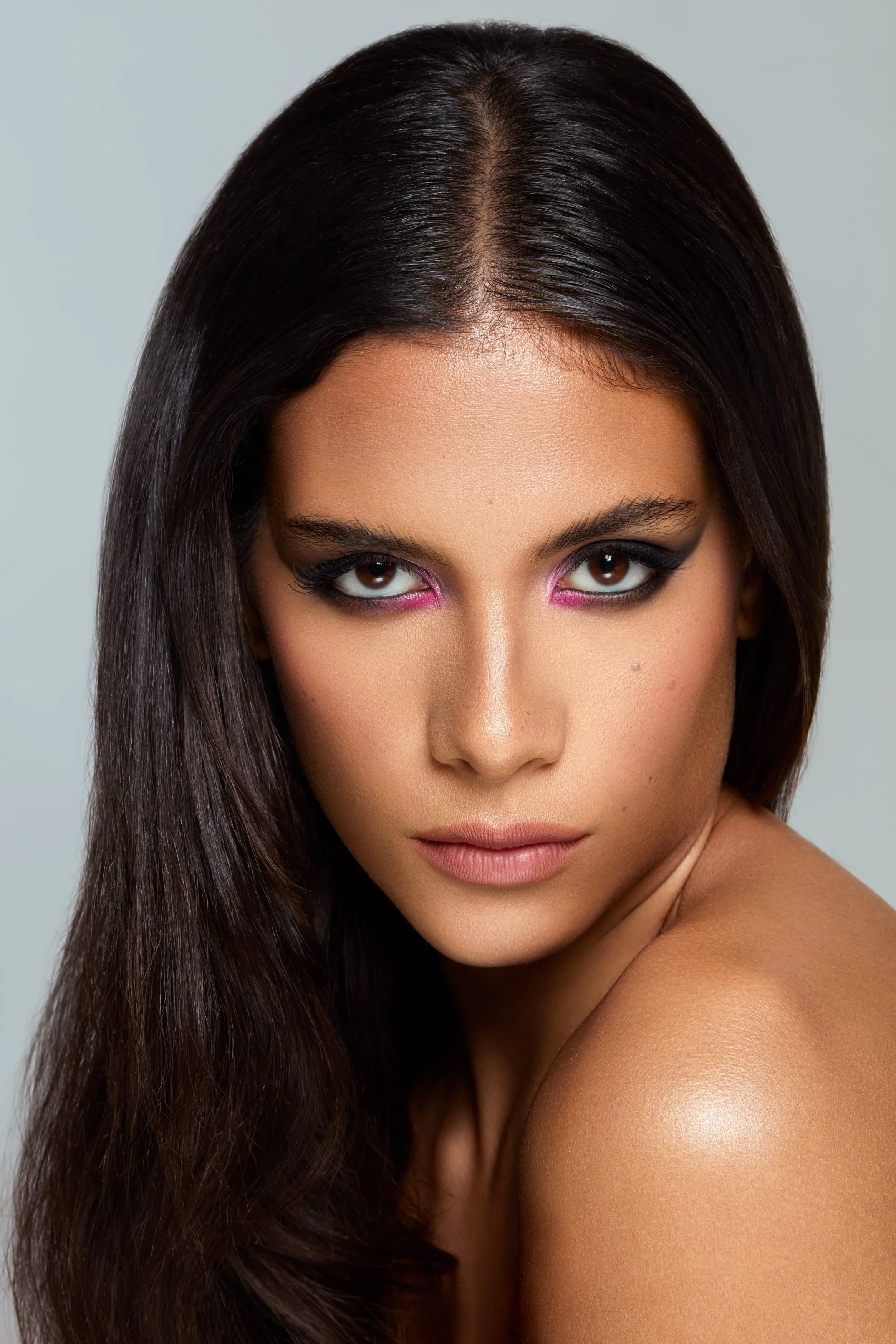 Modella mediterranea mora con occhi scuri indossa makeup intenso smokey eyes con dettagli fucsia accesi, makeup bold e audace luminoso con focus su occhi e spalle in primo piano