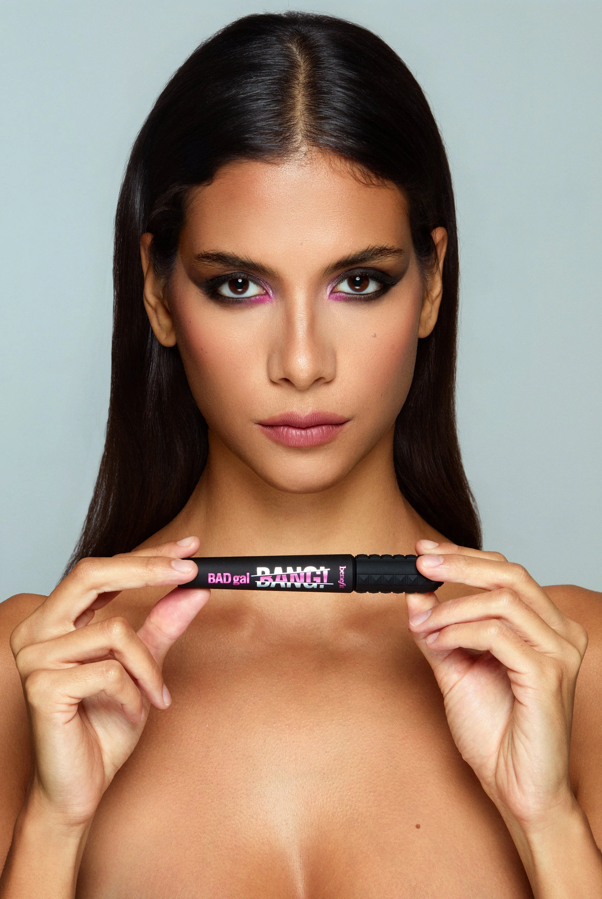 Modella mediterranea capelli neri occhi scuri, capelli lisci dietro le spalle, guarda dritto mostrando tra le mani il mascara bad gal benefit, sguardo intenso sicuro e sensuale, makeup smokey eyes nero con accenti fucsia, pelle naturale luminosa, fot