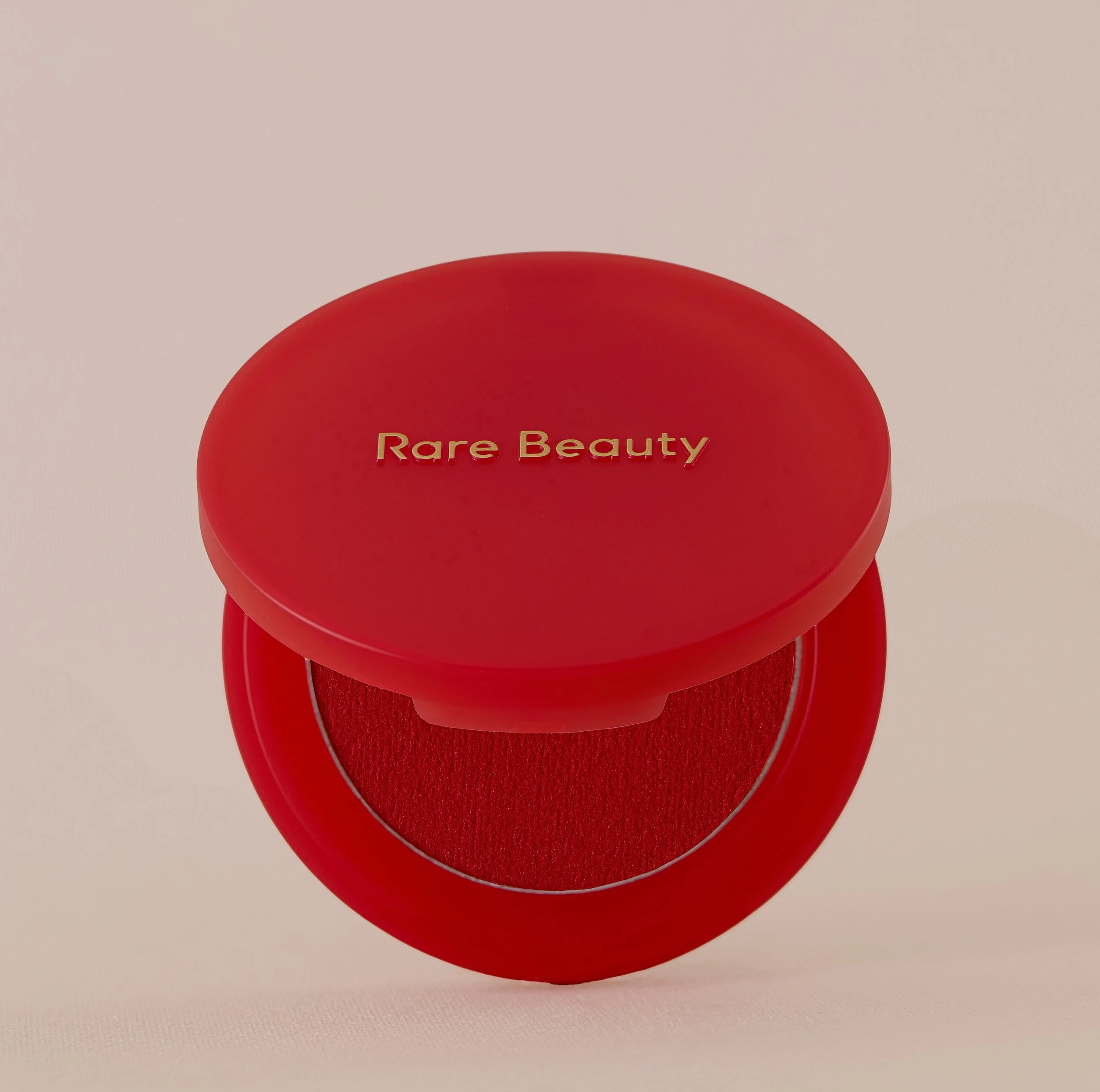 Blush di color rosso acceso rare beauty, poggia su una base beige, lo sfondo tono su tono beige, il prodotto con tappo leggermente aperto in modo da mostrare l'interno del prodotto, di color fucsia acceso, composizione minimal armoniosa che valorizza