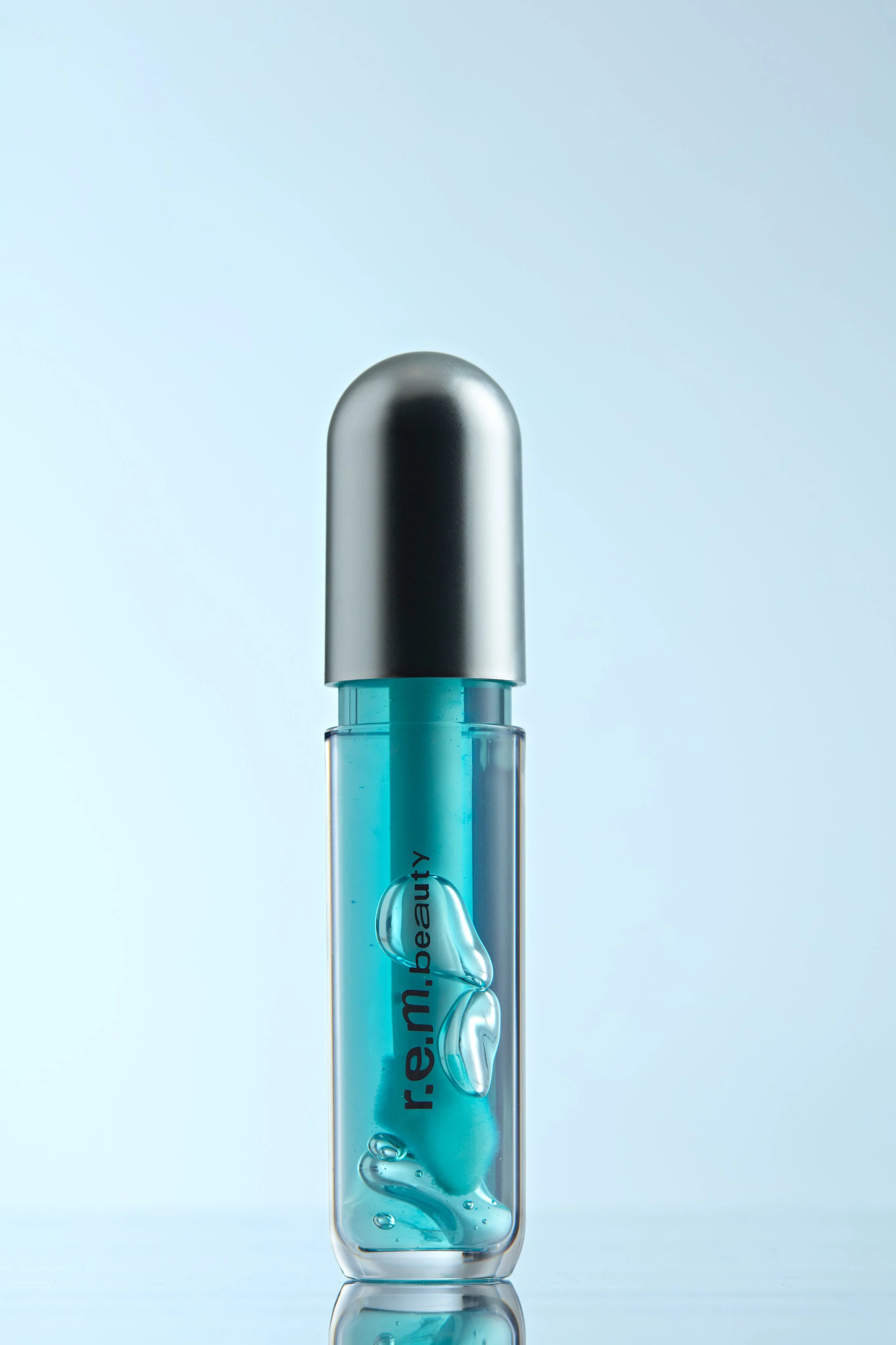 Olio labbra REM beauty, confezione trasparente con all'interno olio labbra color azzurro, poggia su base specchiata, sullo sfondo color ghiaccio, composizione luminosa e moderna che valorizza texture glossy, colore e finish nutriente del prodotto