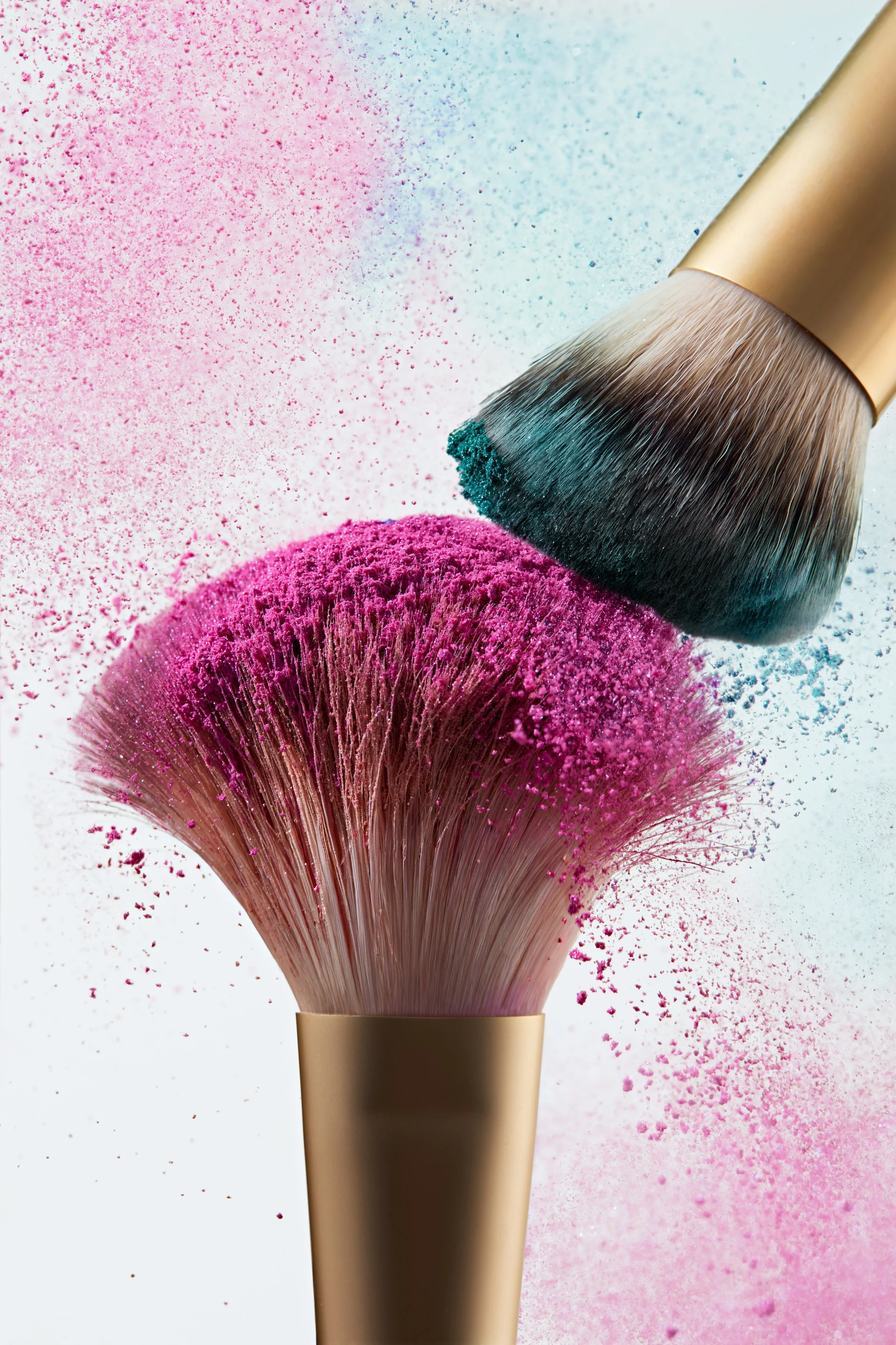 Due pennelli make-up in collisione su sfondo minimal, uno centrale con setole verso l’alto cariche di polvere fucsia e l’altro dall’alto a destra con pigmento blu, con particelle di colore sospese fucsia e blu, che esaltano texture e dinamismo cosmet