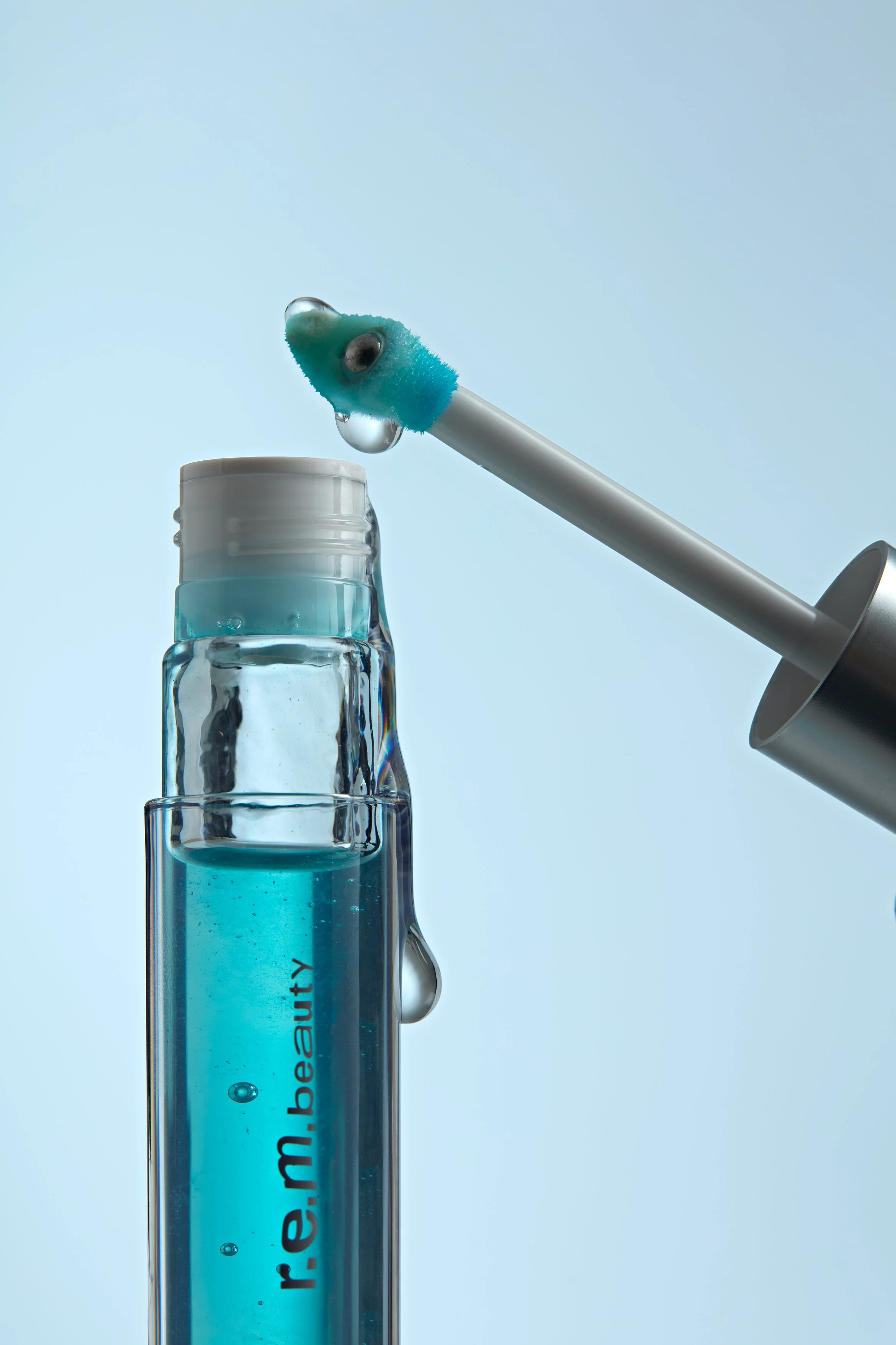 Olio labbra REM beauty, confezione trasparente con all'interno olio labbra color azzurro, su sfondo color ghiaccio, l'applicatore è sospeso, dalla confezione e dall'applicatore cola il prodotto trasparente, composizione moderna che valorizza texture 