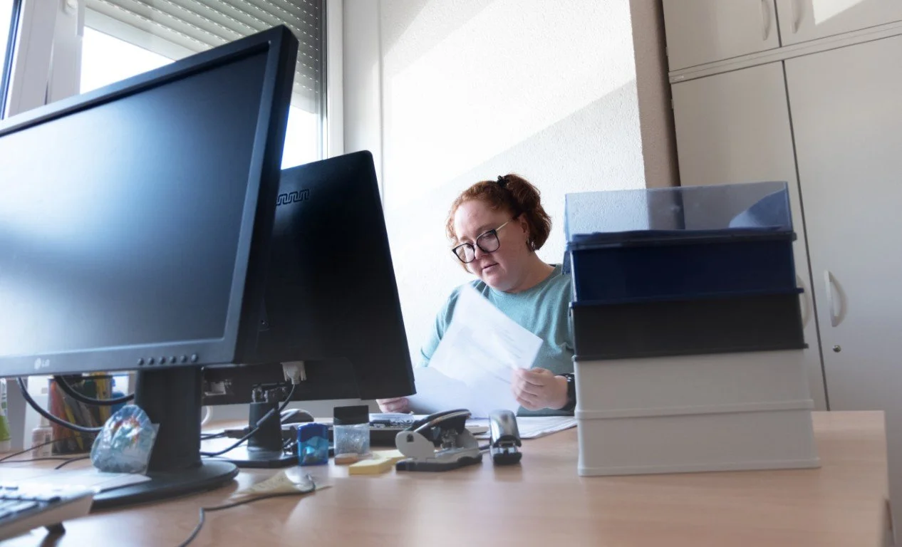 Frau sitzt an einem Schreibtisch in einem Büro und liest Dokumente. Im Vordergrund sind zwei Computermonitore, Bürobedarf und Aktenordner sichtbar.