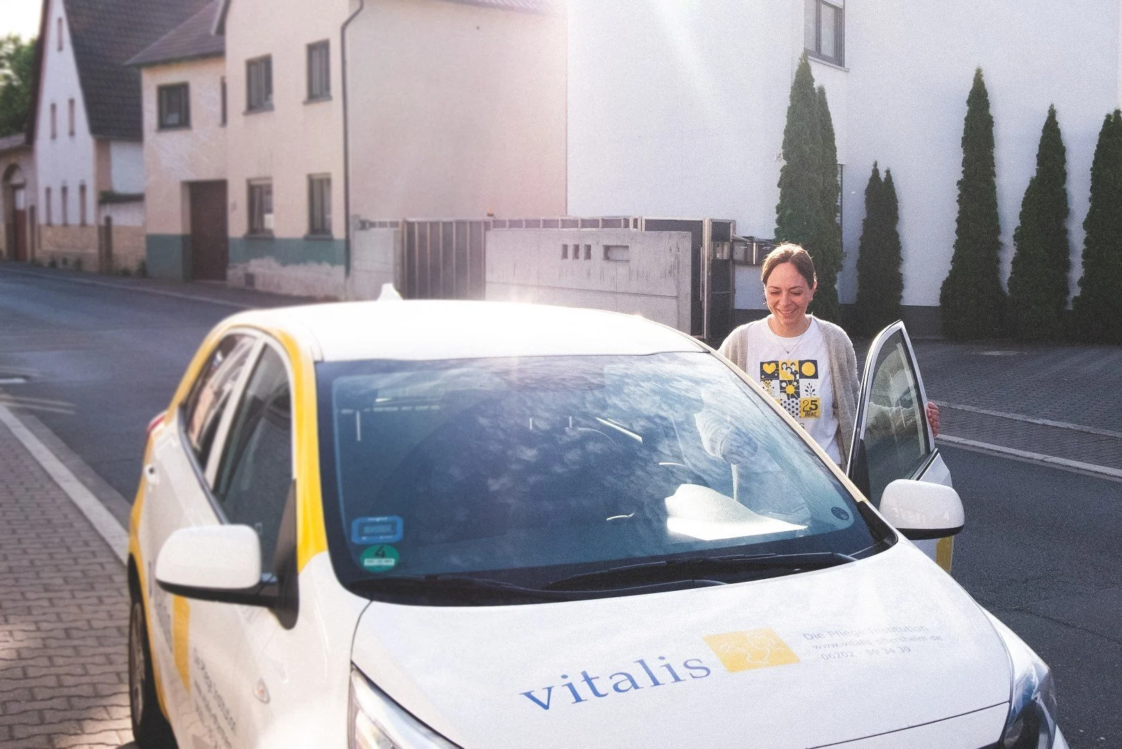 Eine lächelnde Frau öffnet die Fahrertür eines weißen Autos mit der Aufschrift 'Vitalis' vor einem Wohnhaus auf einer ruhigen Straße.