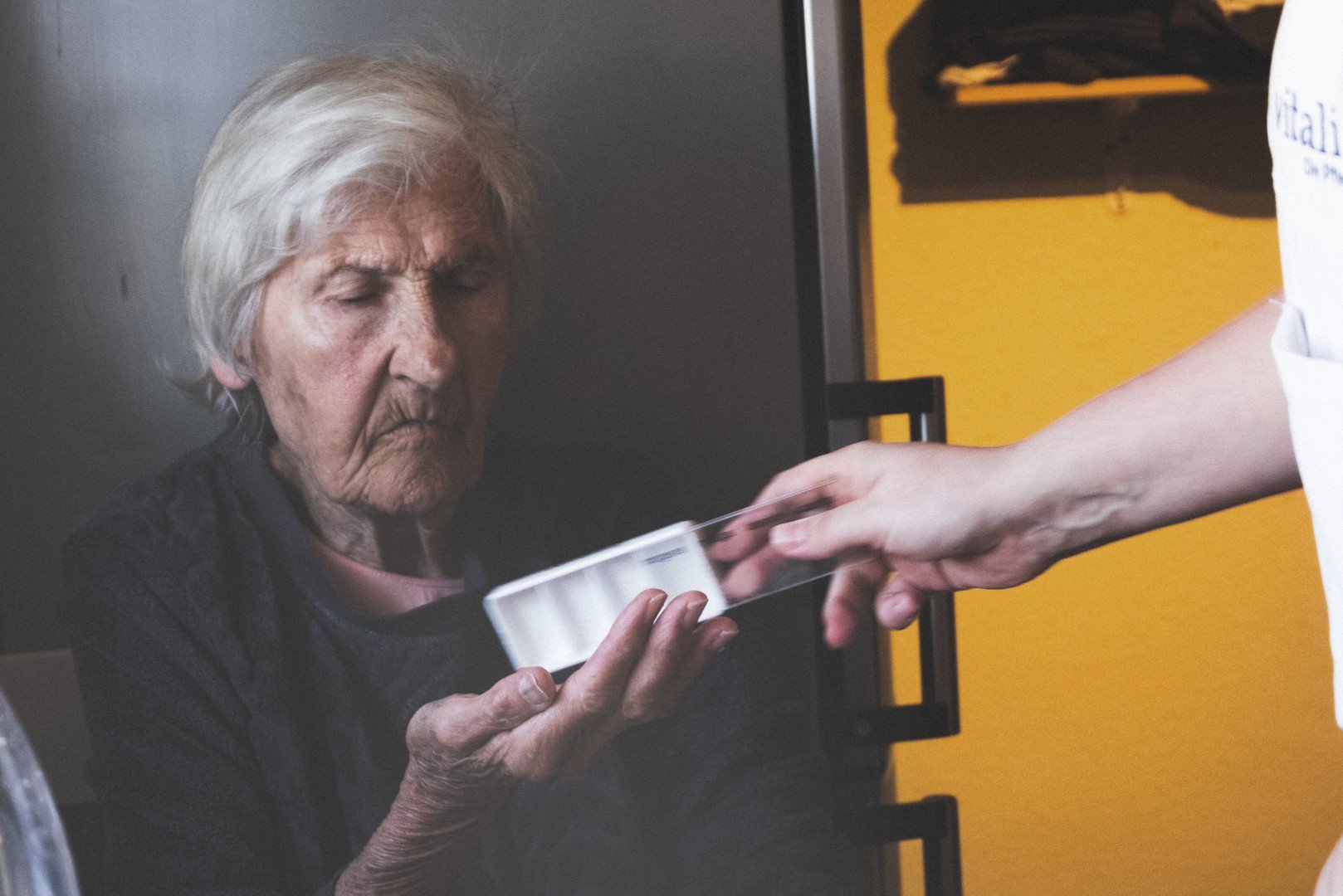 Eine ältere Frau mit weißen Haaren erhält ein Medikament in einer kleinen Verpackung von einer helfenden Person, während sie mit geschlossenen Augen blickt.