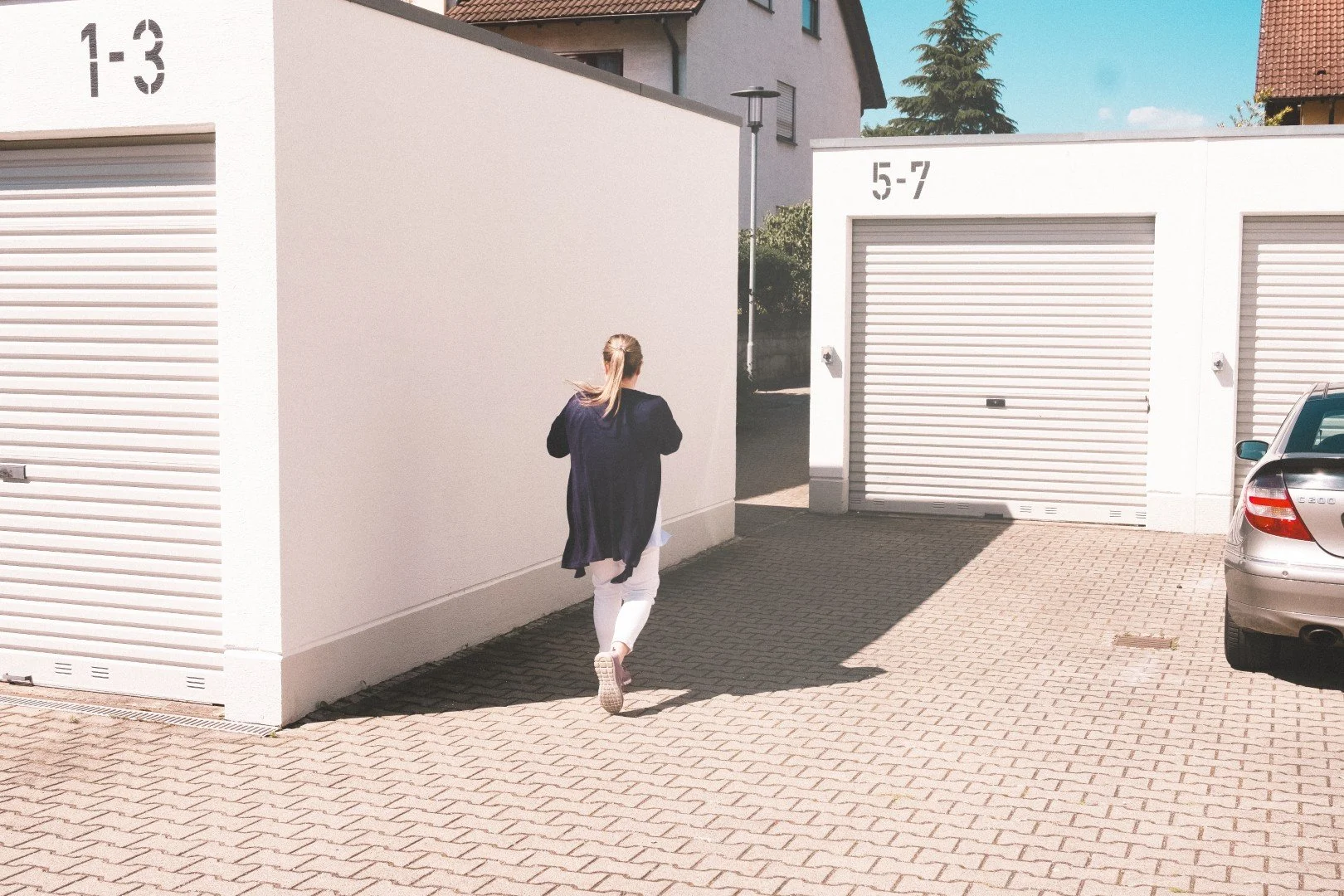 Eine Frau läuft auf einer gepflasterten Zufahrt neben einem weißen Gebäude mit Garage-Doors. Es ist sonnig und der Himmel ist blau.