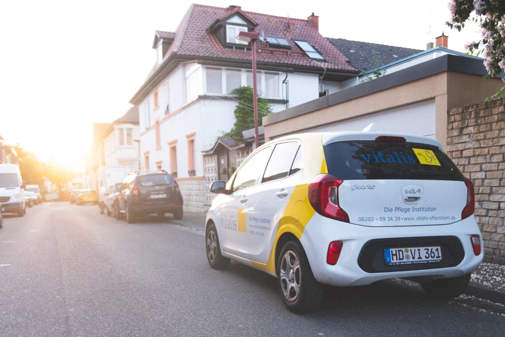 Ein kleines Auto mit der Werbung für Vitalis Pflegeinstitut geparkt an einer ruhigen Wohnstraße bei Sonnenuntergang, mit mehreren parkenden Fahrzeugen entlang der Straße und einem weißen Haus im Hintergrund.