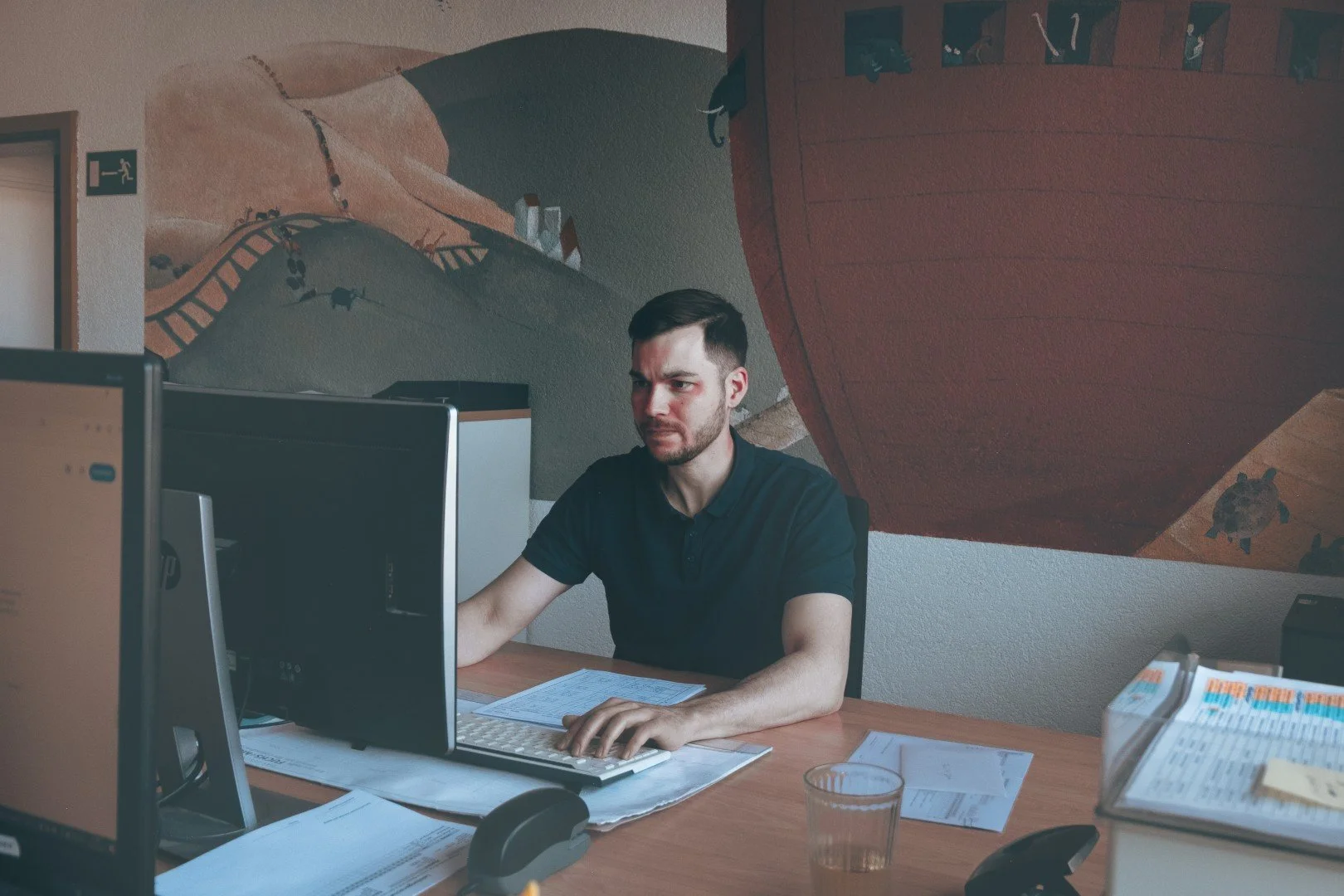 Junge Mann arbeitet an einem Schreibtisch im Büro, umgeben von Papieren, Computern und einem Telefon, hinter einer Wand mit einer Gemäldewand.