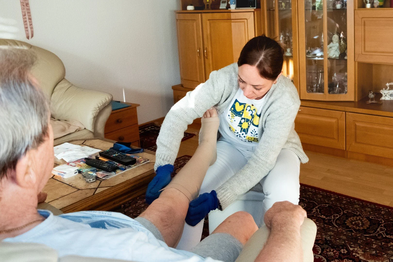 Frau und älterer Mann in einer Wohnung, Frau hilft dem Mann beim An- oder Ausziehen einer elastischen Unterstützung für das Bein, während sie auf einem Stuhl sitzt.