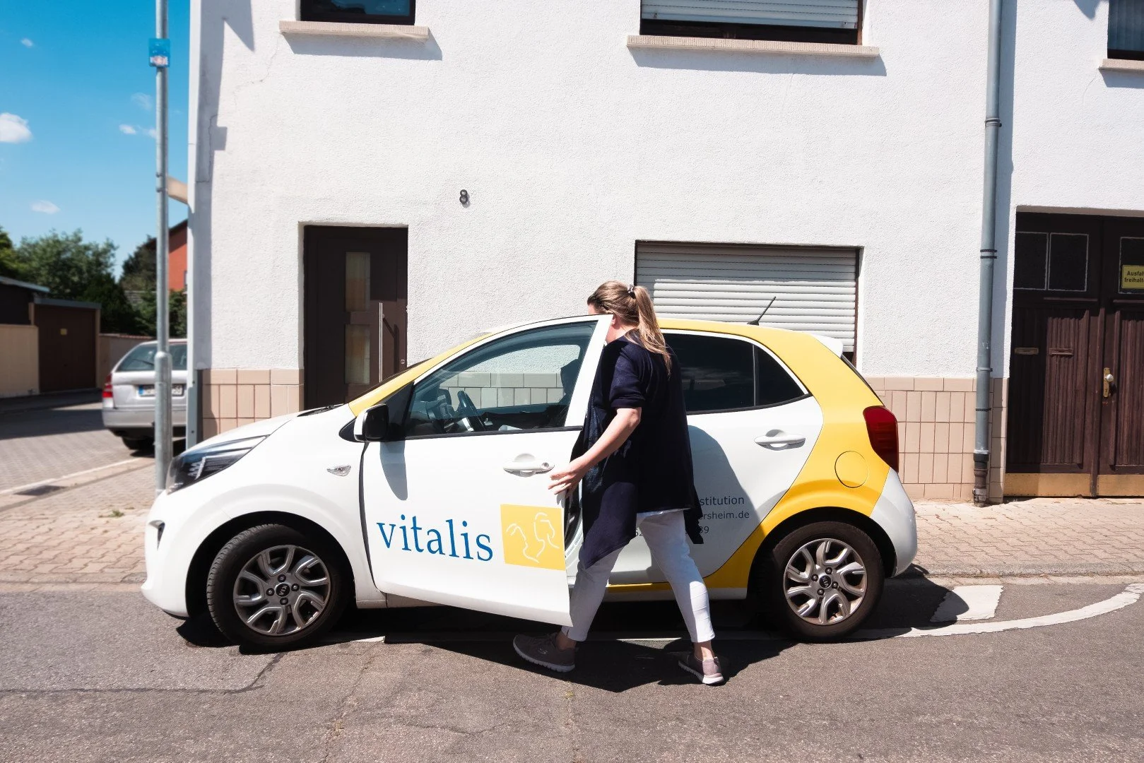 Frau steigt in ein weiß-gelbes Auto mit der Aufschrift 'Vitalis', das an einer Straßenrandparken. Das Auto ist vor einem weißen Gebäude mit geschlossenem Fensterladen geparkt an einem sonnigen Tag.