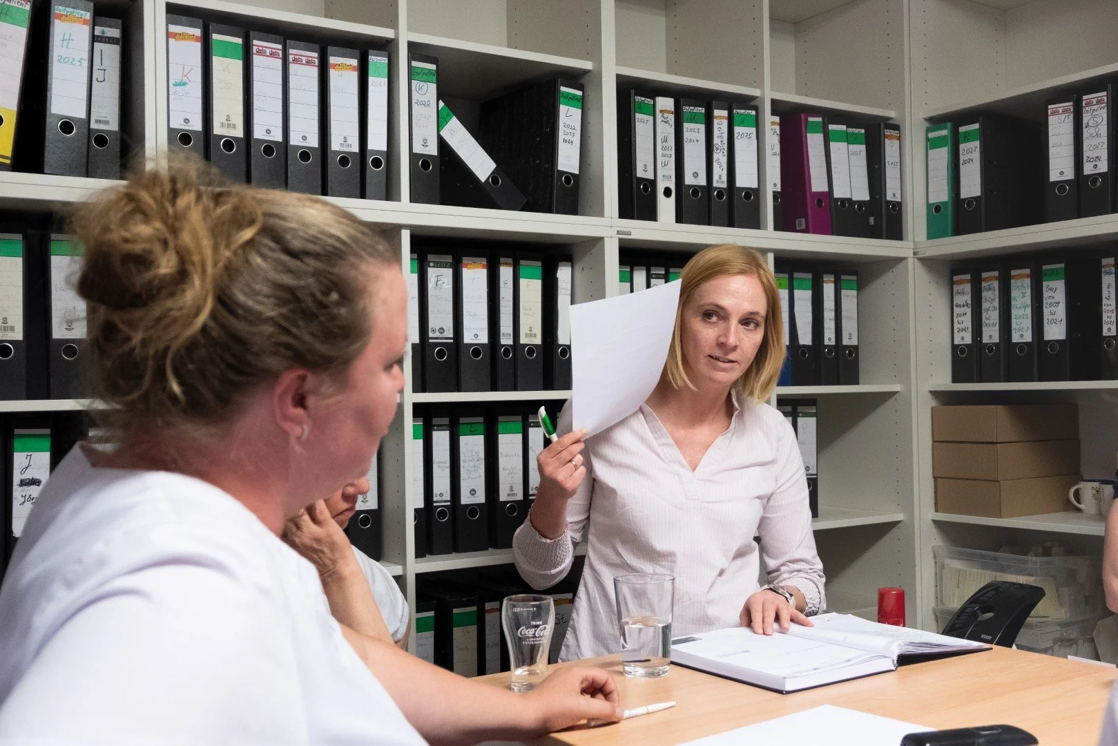 Drei Frauen in einem Büro bei einer Besprechung. Eine Frau hält ein Blatt Papier in der Hand und spricht, die anderen hören zu.