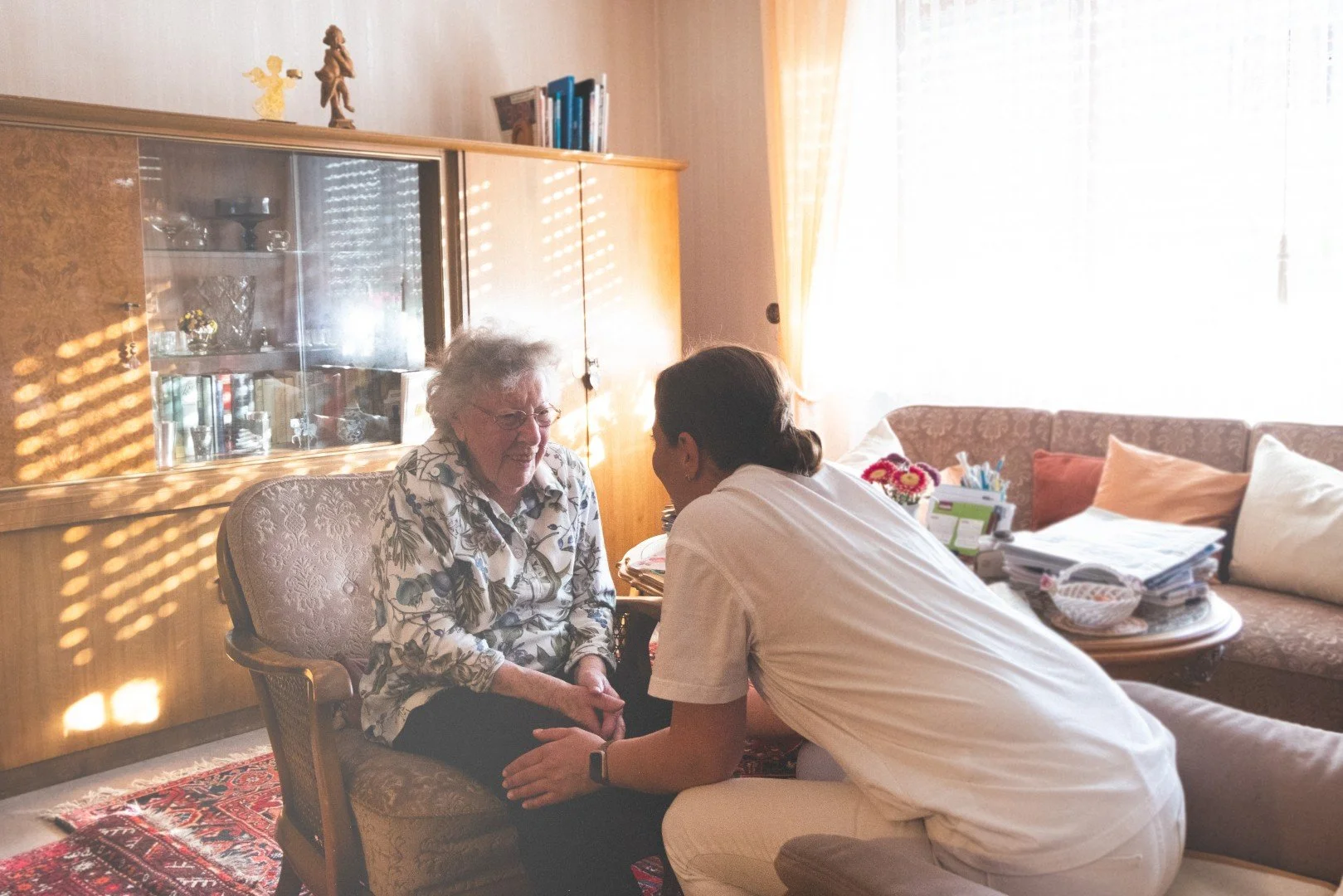 Eine Pflegekraft spricht mit einer älteren Frau in einem Wohnzimmer, Sitzecke mit Couch und viel Tageslicht im Hintergrund.