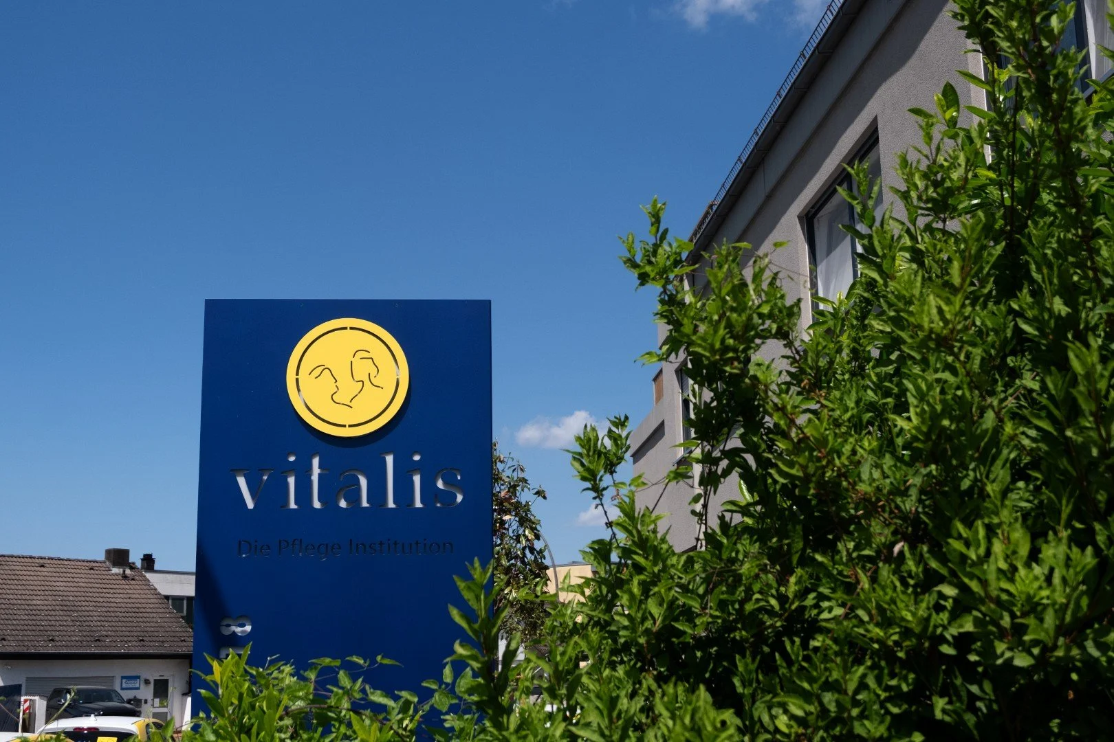 Blaue Werbetafel für die Pflegeeinrichtung Vitalis mit einem Logo, umgeben von grünen Sträuchern, vor einem Gebäude mit weißen Wänden und mehreren Fenstern, bei sonnigem Himmel.